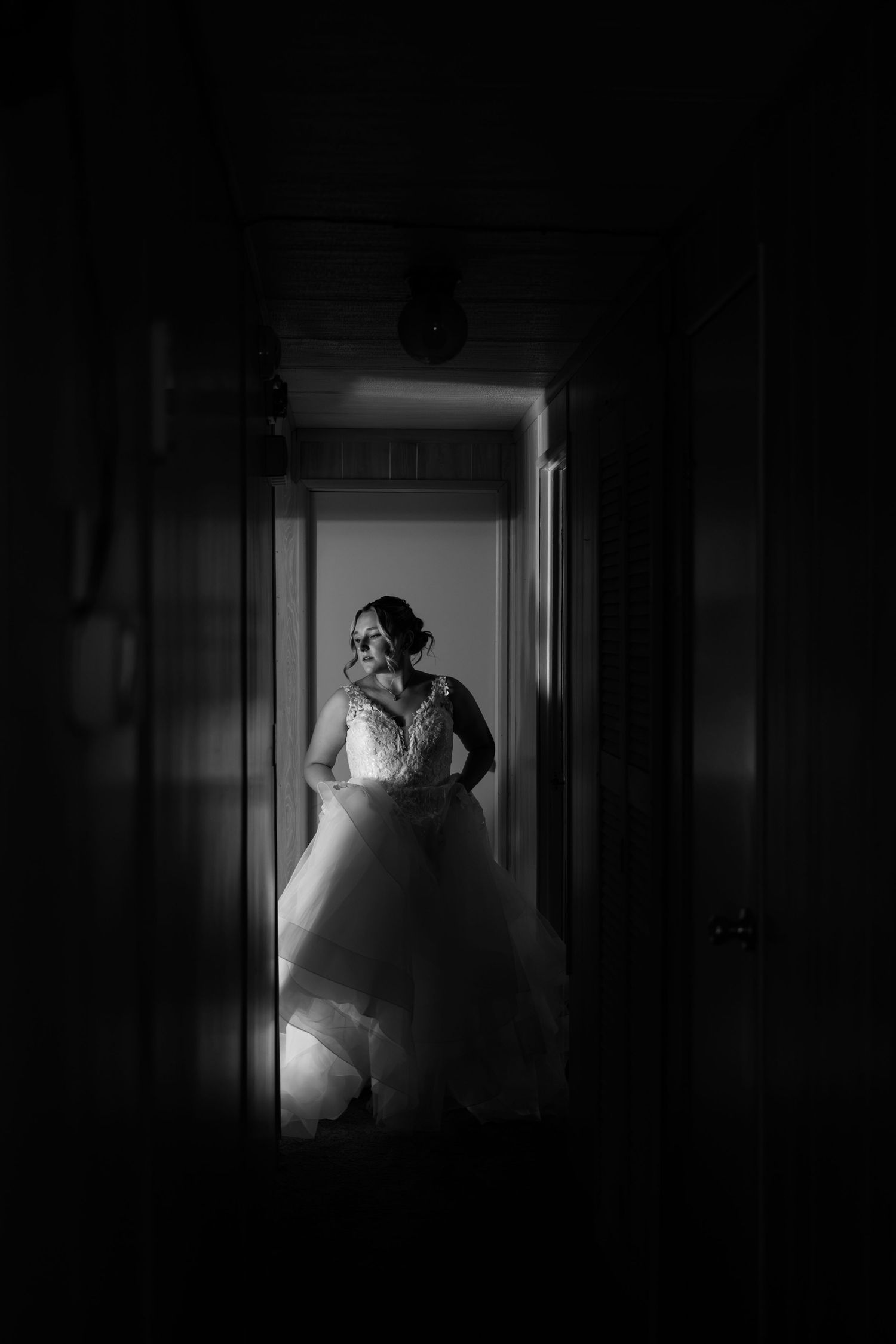 Dramatic black and white portrait of a bride in a ballgown standing in a dimly lit hallway.