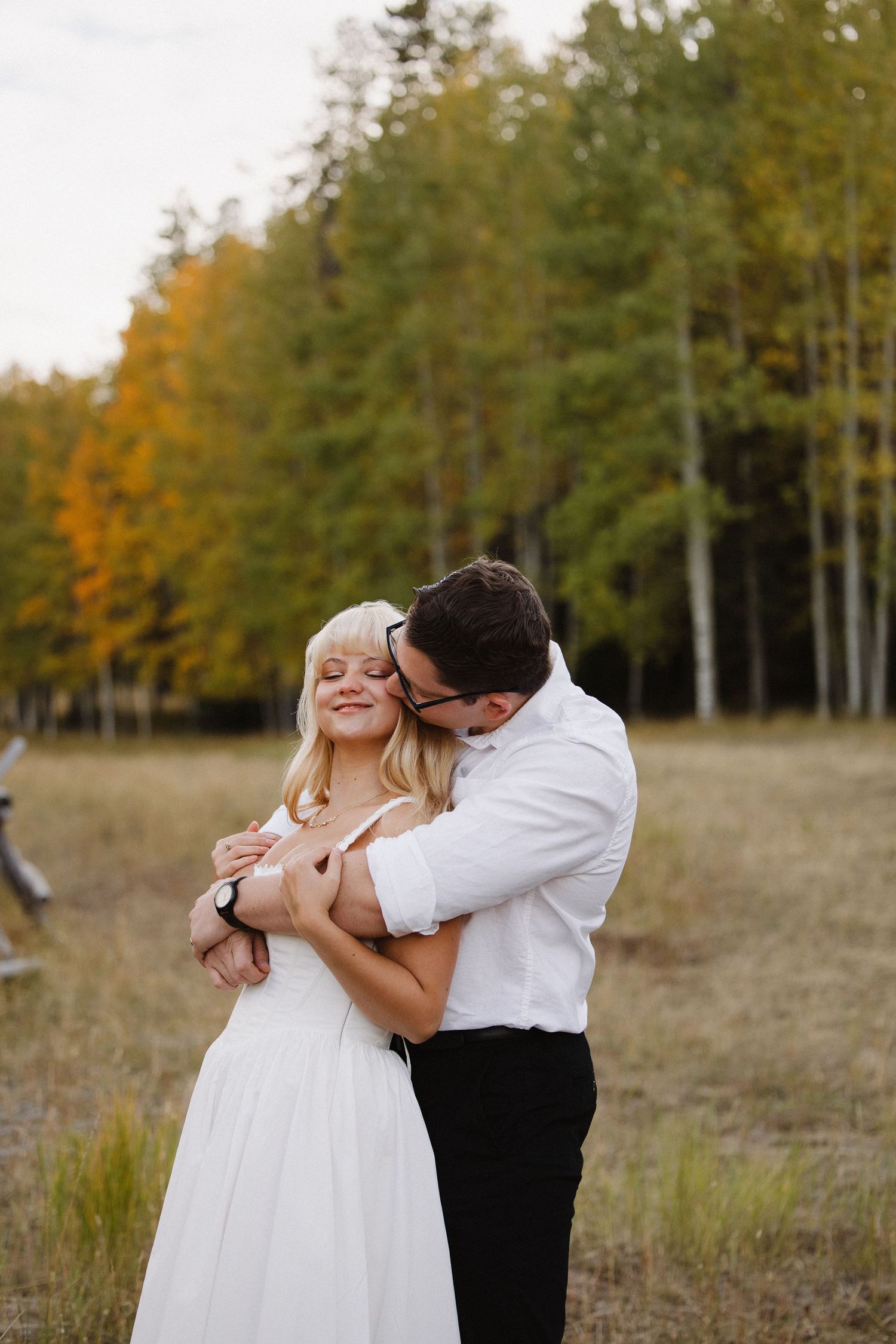 Groom in white shirt embraces smiling bride from behind amid colorful fall trees in open field. Wyoming wedding.