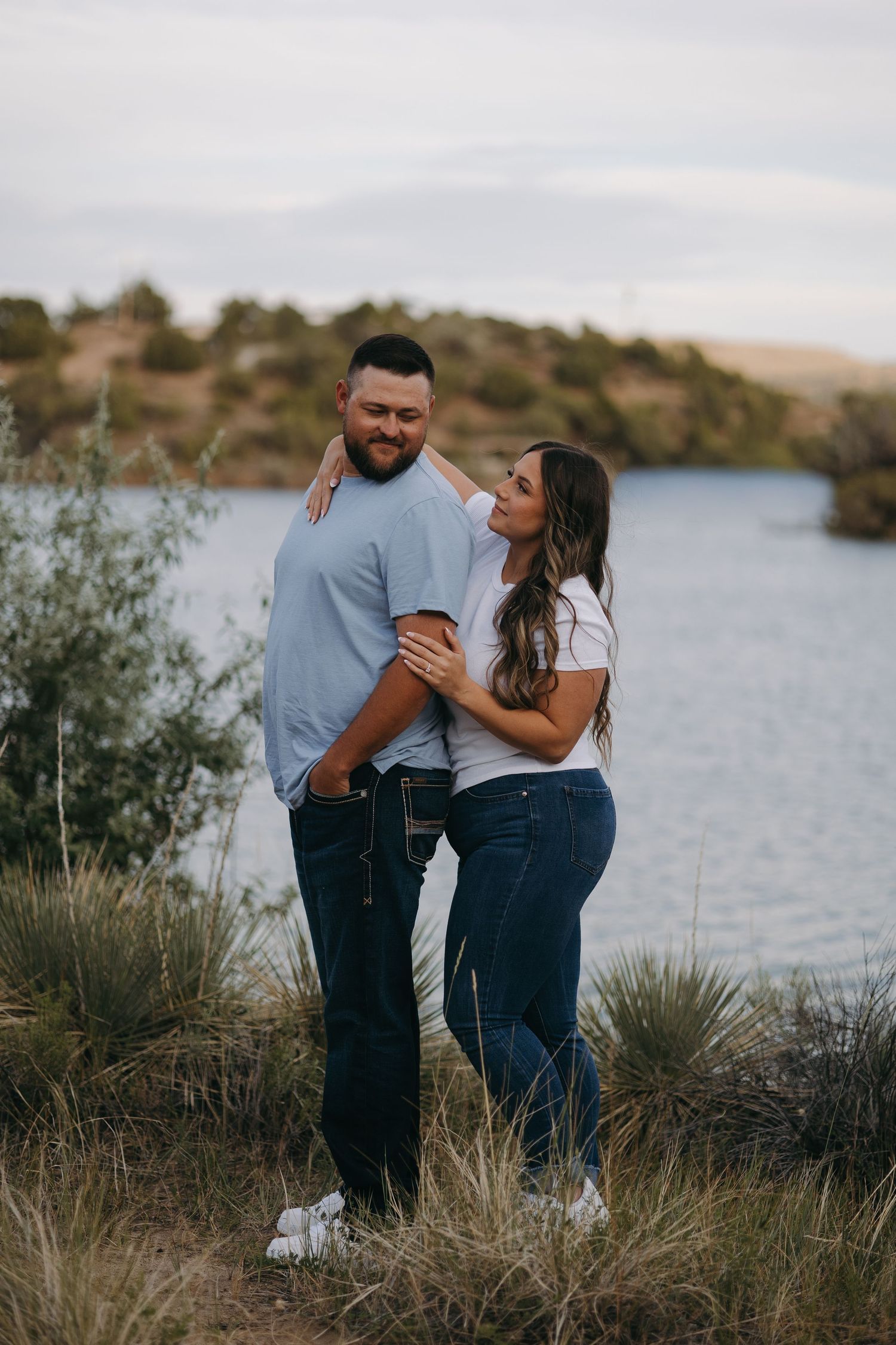 Couple poses together near a lake at dusk, woman embracing man from behind in casual attire.