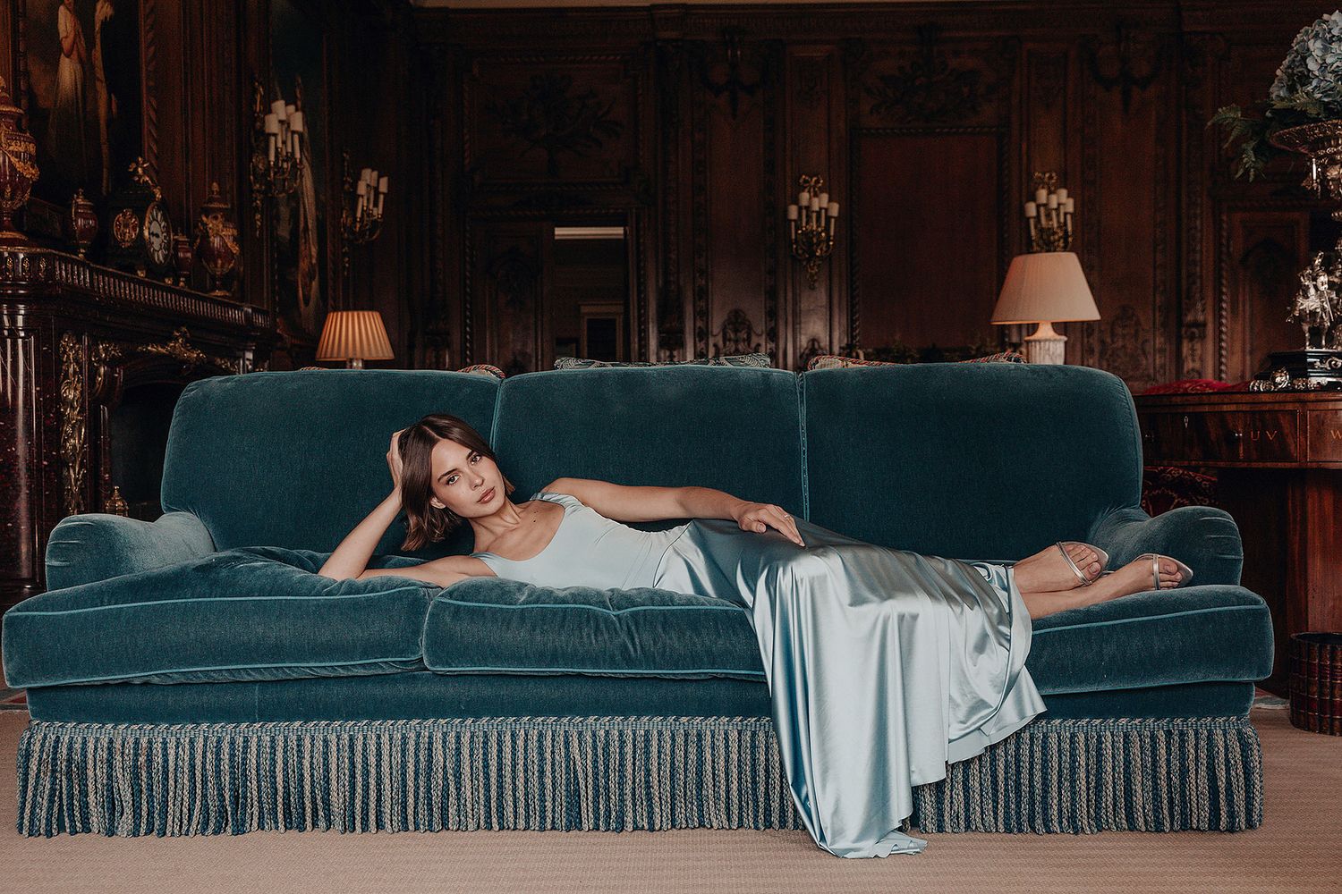 Woman in a silky pale blue slip dress reclining on a green velvet sofa in an elegant wood-paneled room.
