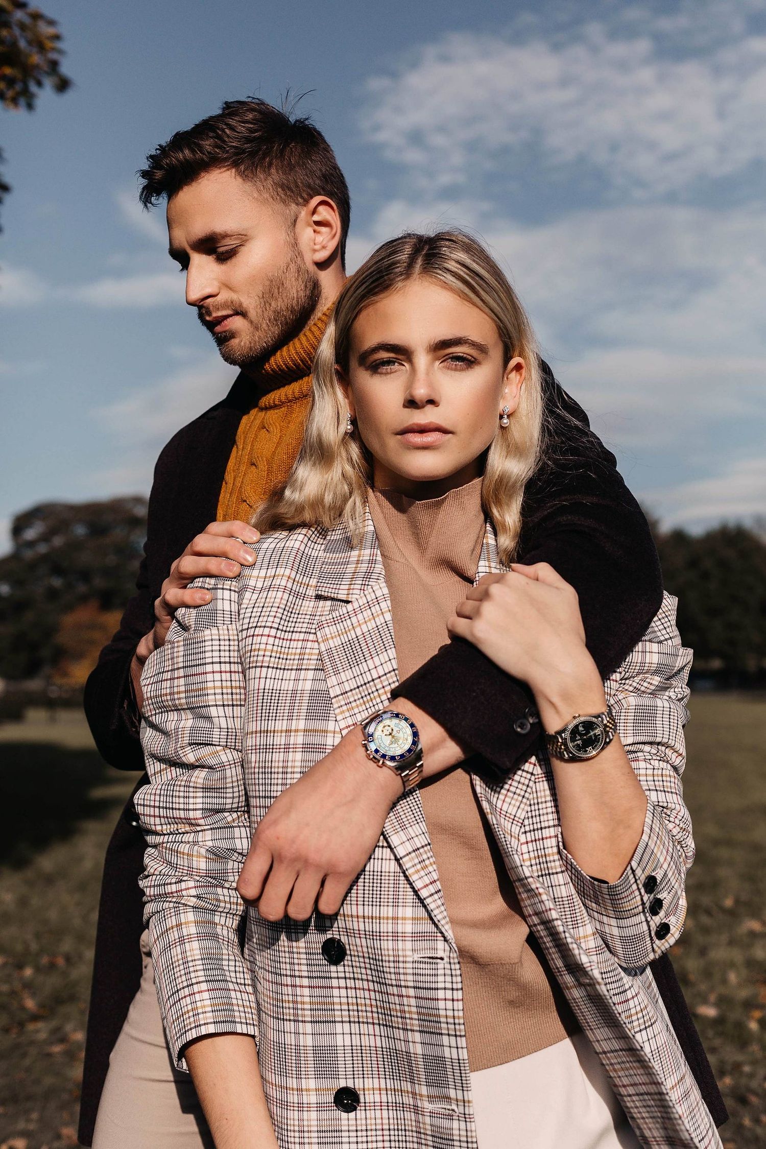 Couple embracing outdoors under a blue sky, she in a plaid blazer with wristwatches, he in a dark coat and mustard turtleneck.