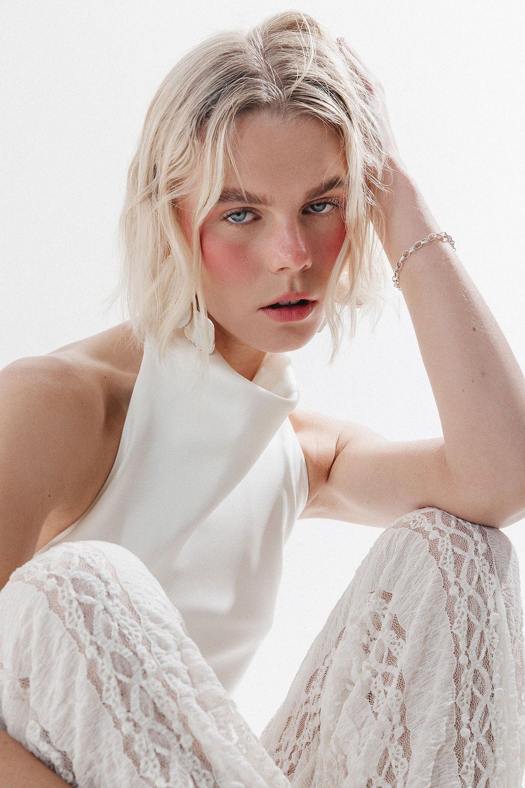 Blonde woman with short wavy hair and rosy cheeks wears a white halter top and lace pants, hand resting on her head.