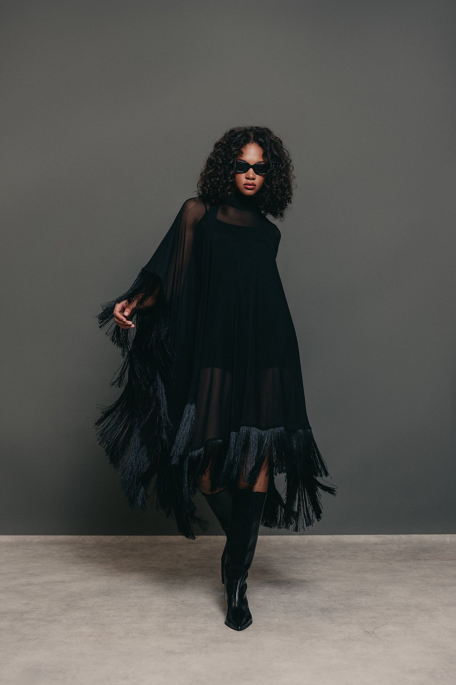 Woman in sunglasses and a flowing black fringed chiffon dress with tall black boots posing against a gray backdrop.