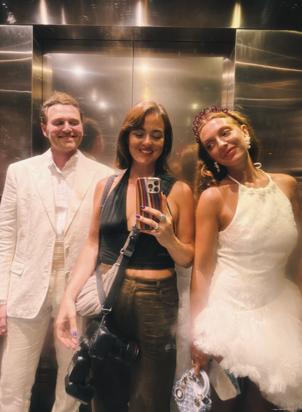 Three smiling friends in elegant white and black outfits pose for a mirror selfie inside a metallic elevator.