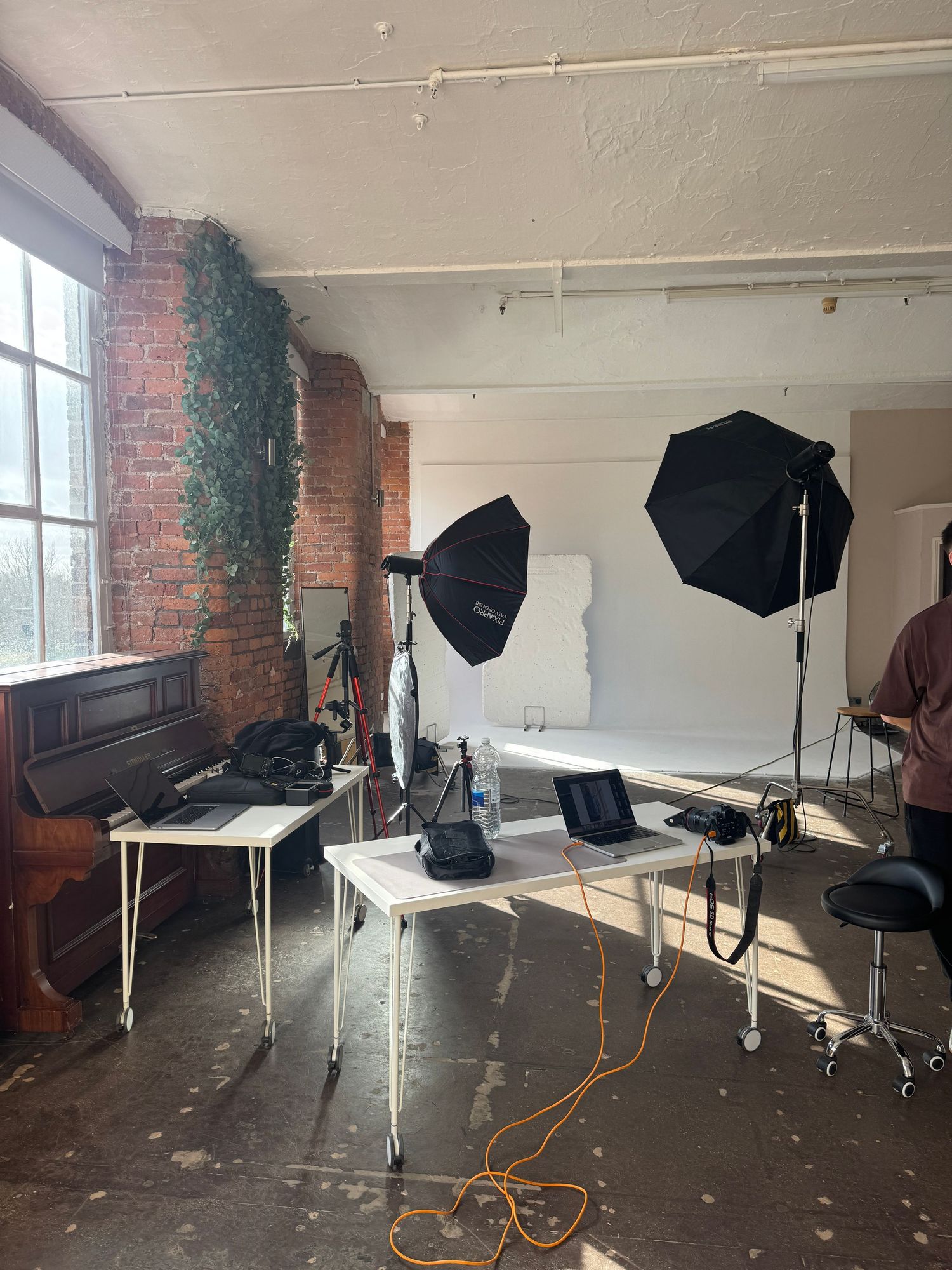 Photography studio with brick walls, softbox lights, tripods and a laptop on tables beside a vintage upright piano.