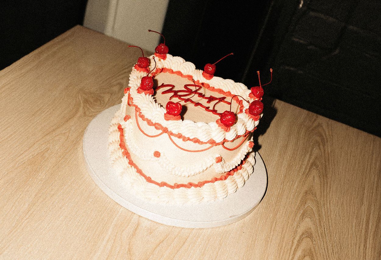 Retro heart-shaped cake decorated with red icing details and cherries, displayed on a white board on a wooden table.