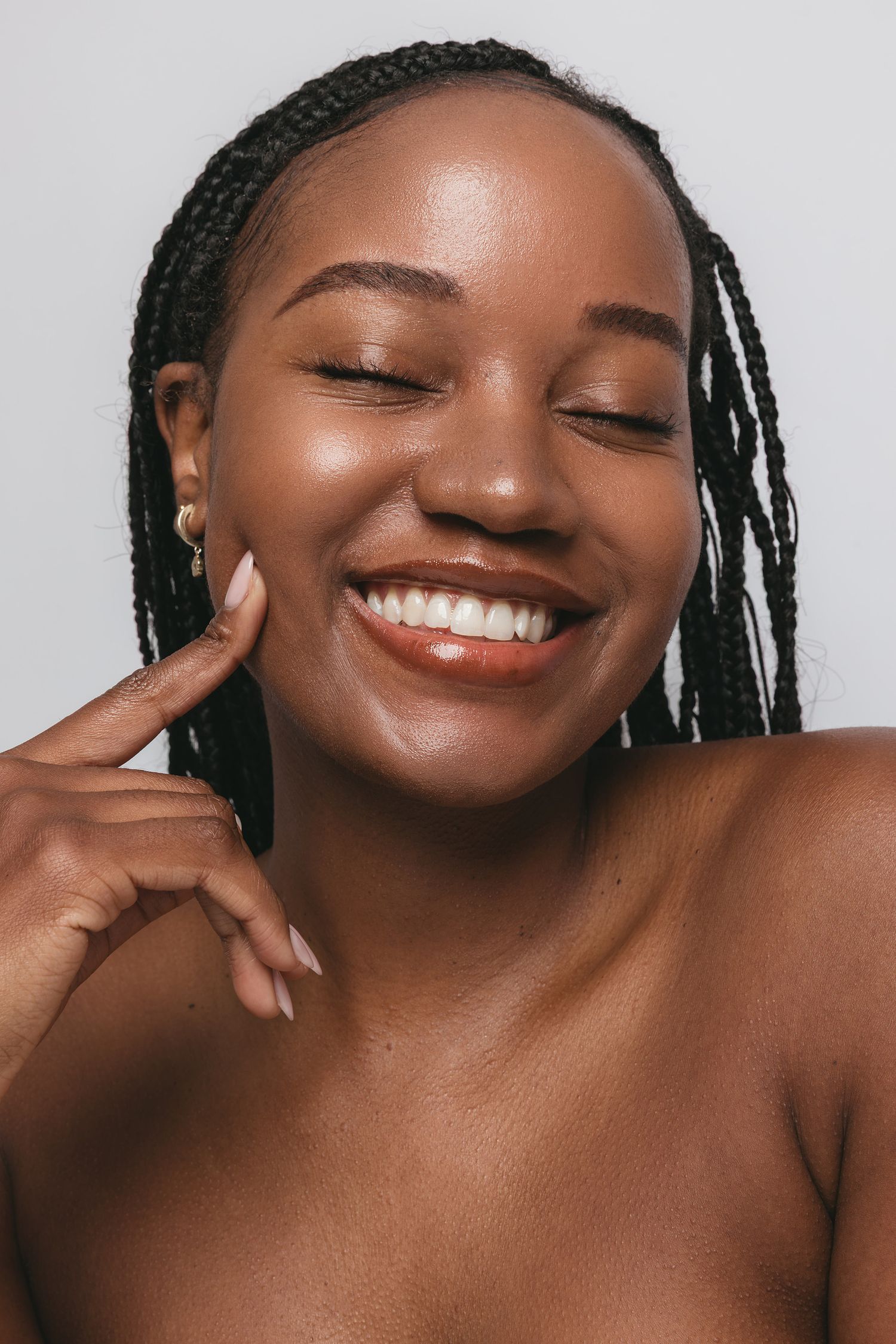 Smiling woman with closed eyes and long braids touches her cheek, showing glowing skin in a bright beauty portrait.