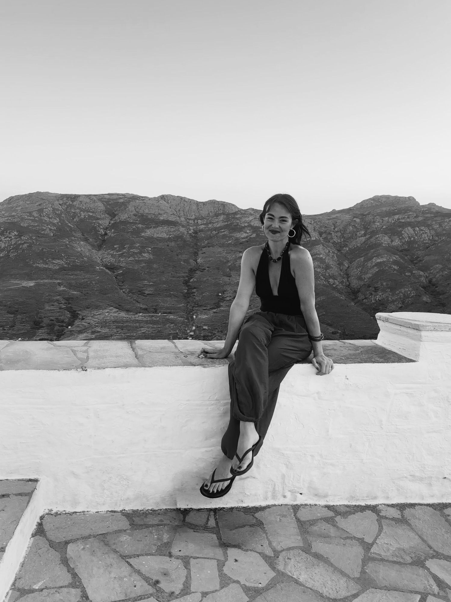 Black and white photo of a smiling woman in a halter top and flip-flops sitting on a whitewashed wall with rugged mountains behind her.