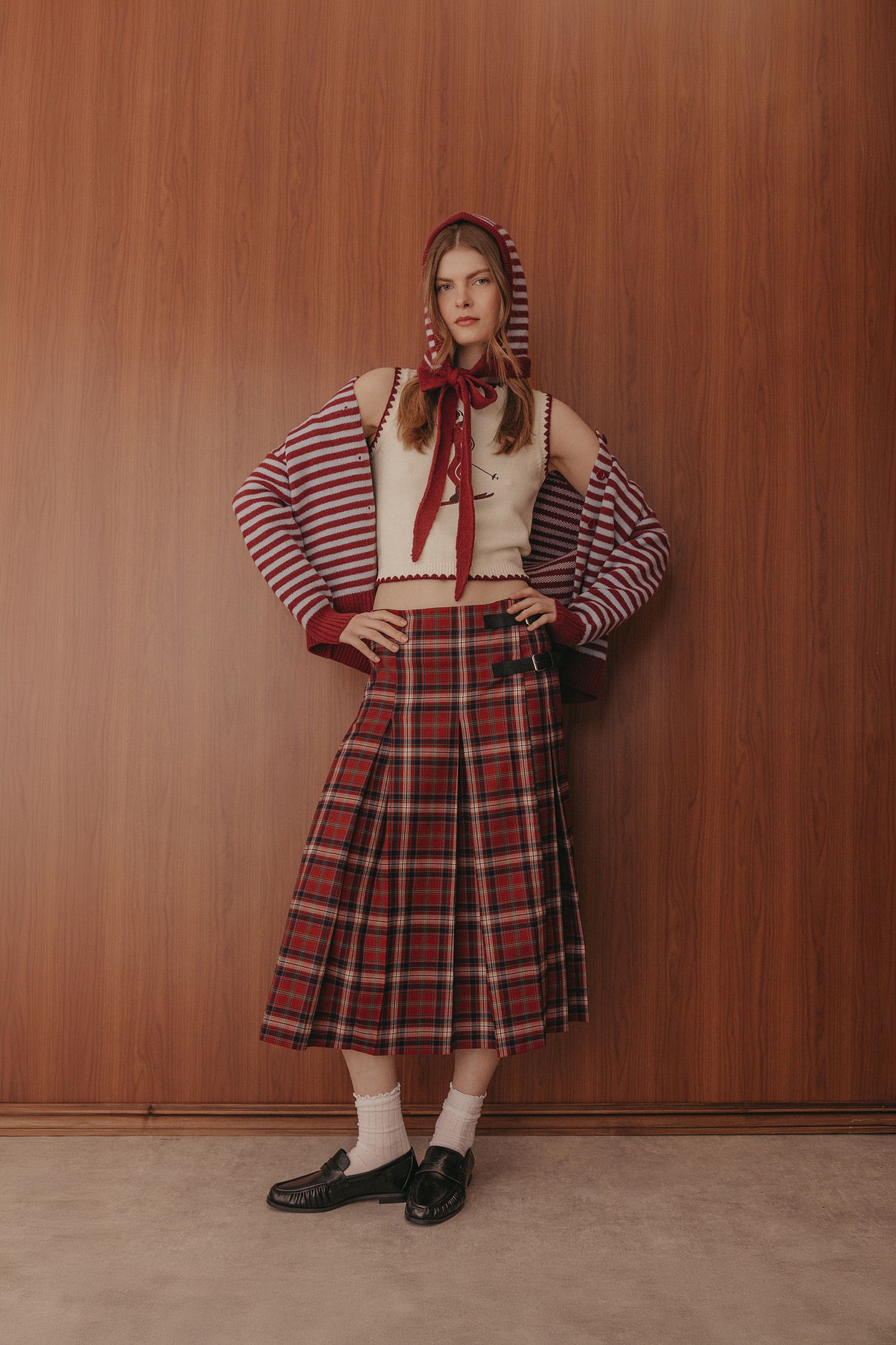 Model in a red plaid pleated skirt, striped cardigan and red hooded scarf poses against a warm wood-paneled wall.