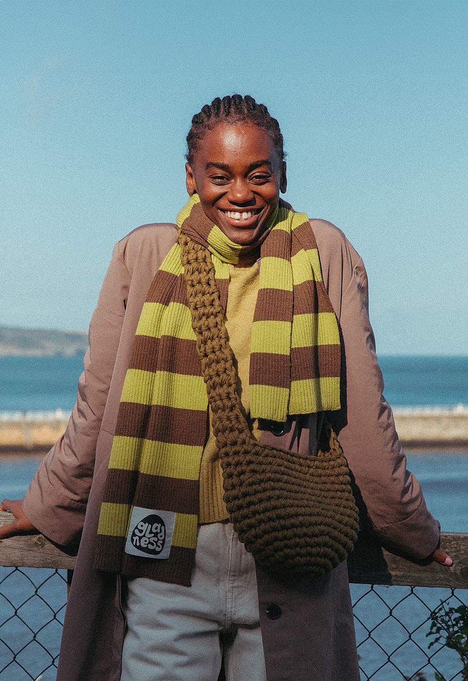 Smiling woman in a yellow and brown striped scarf, olive crochet bag and tan coat leaning on a railing by the sea.