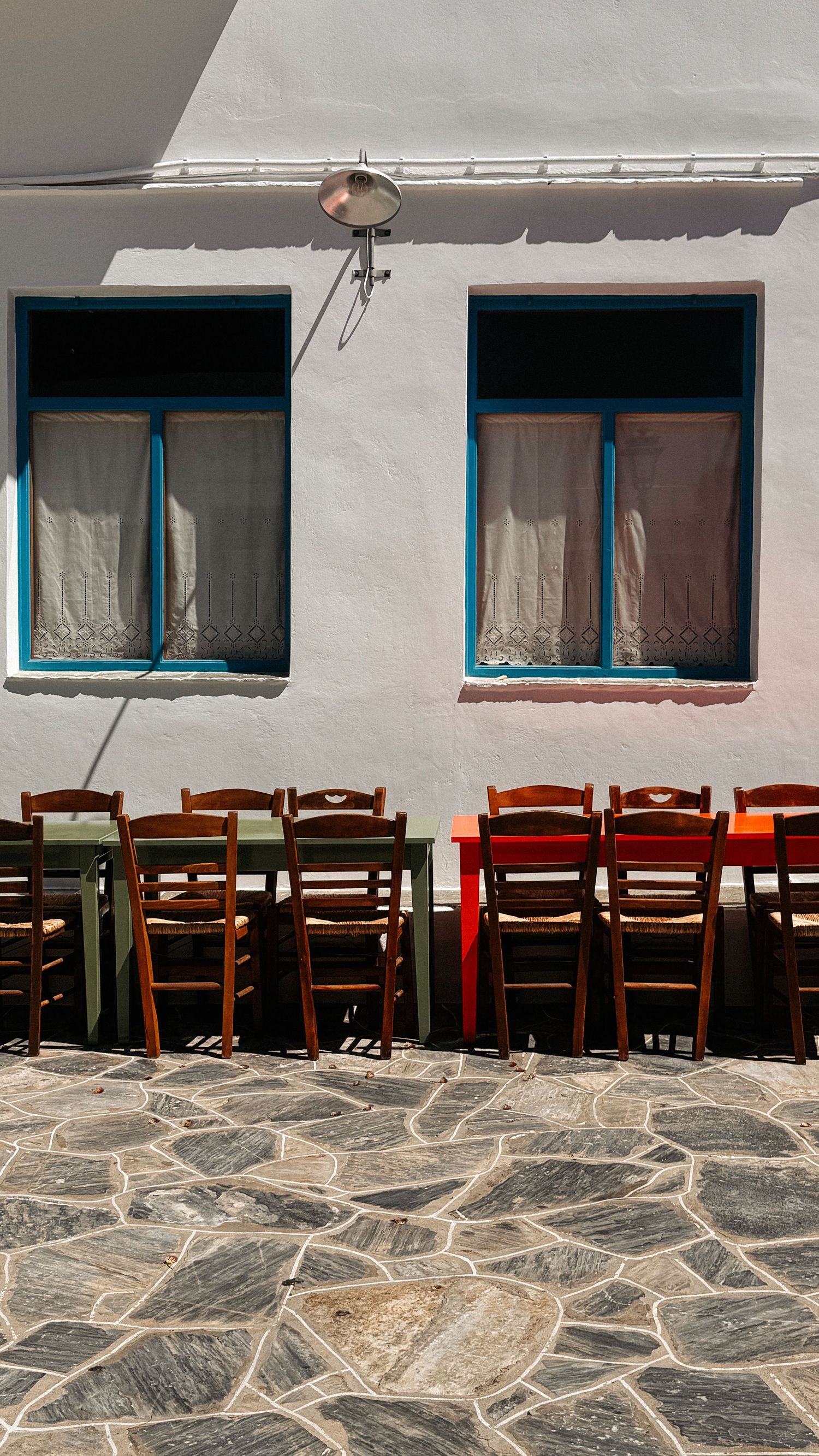 Wooden chairs and colorful tables line a whitewashed wall with blue-framed windows on a stone-paved square.
