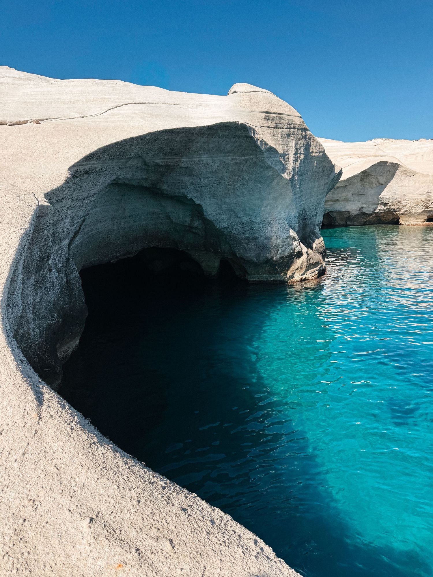 Smooth white volcanic rock formations frame a sea cave with vivid turquoise water under a clear blue sky.