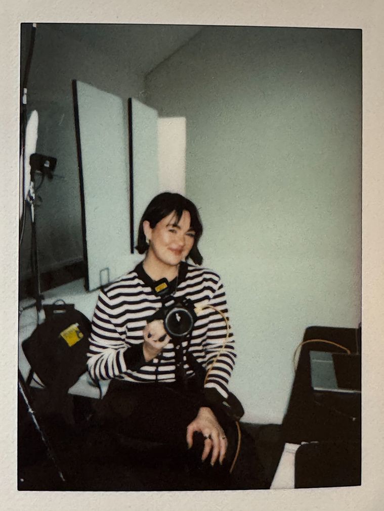 A smiling woman in a striped sweater holds a camera while seated in a photo studio in this instant film portrait.
