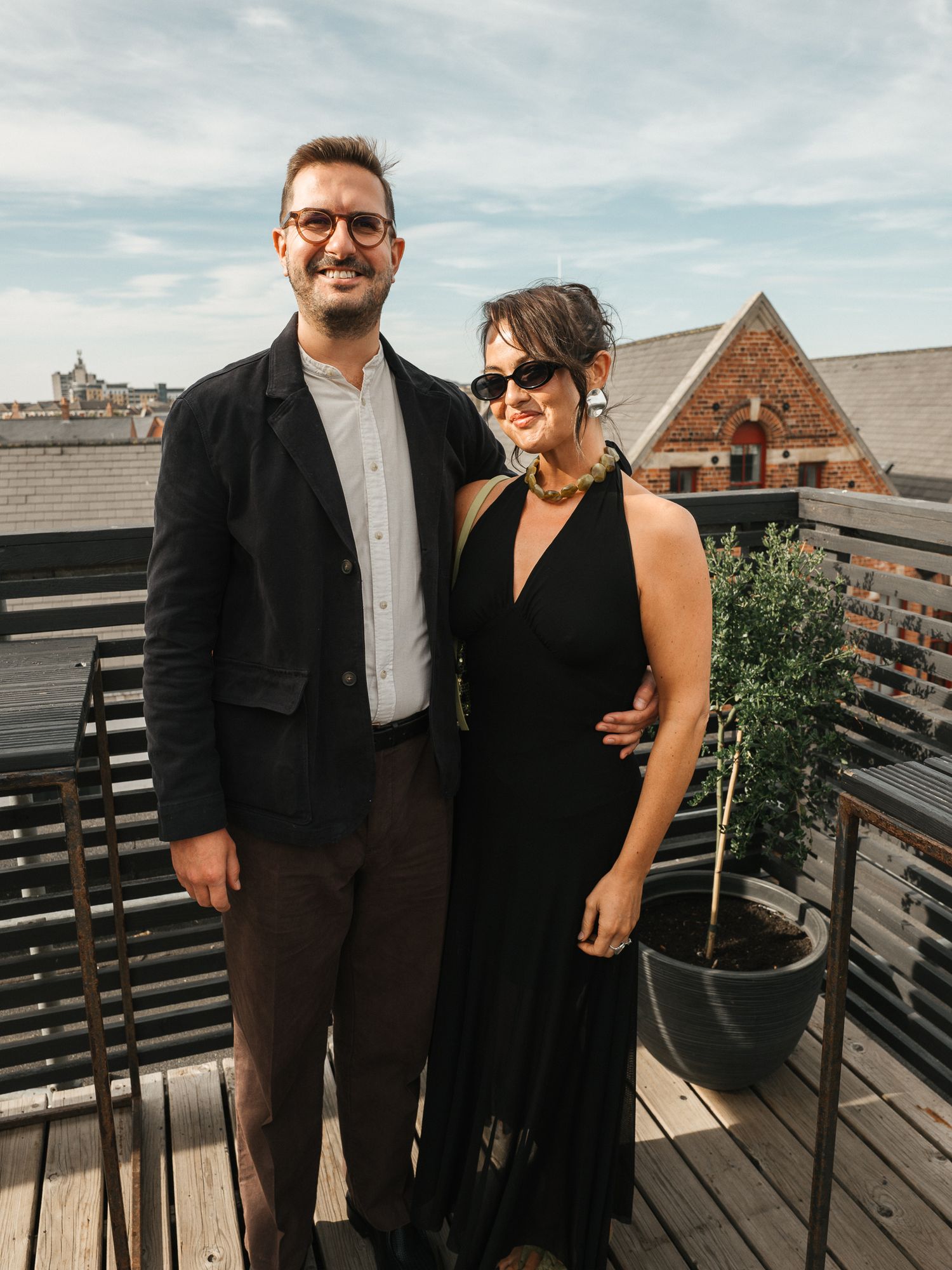 Smiling man in glasses and dark jacket stands beside a woman in a black dress and sunglasses on a rooftop terrace.