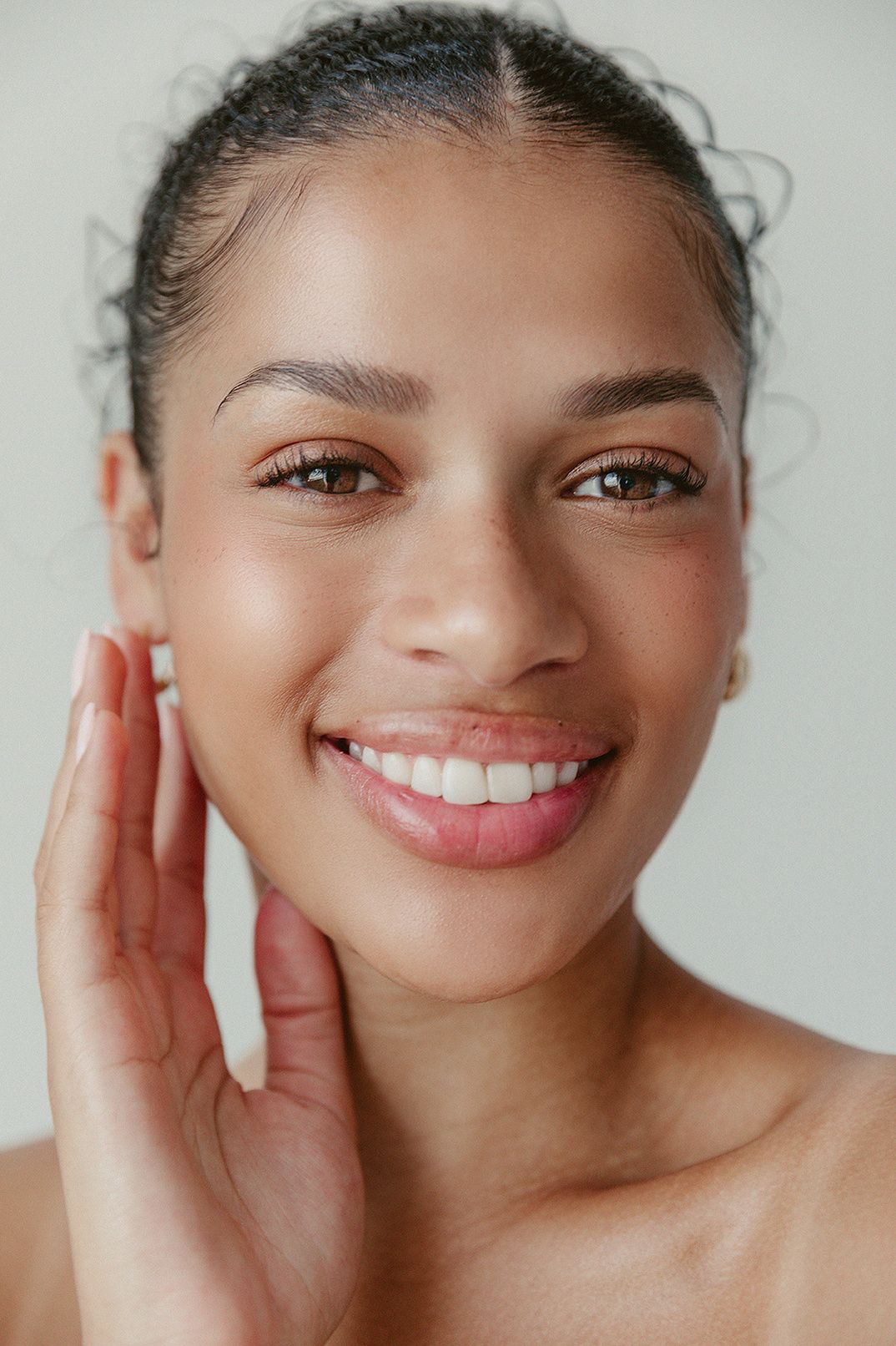 Close-up portrait of a smiling woman with glowing skin and soft pink lips, one hand gently touching her face.