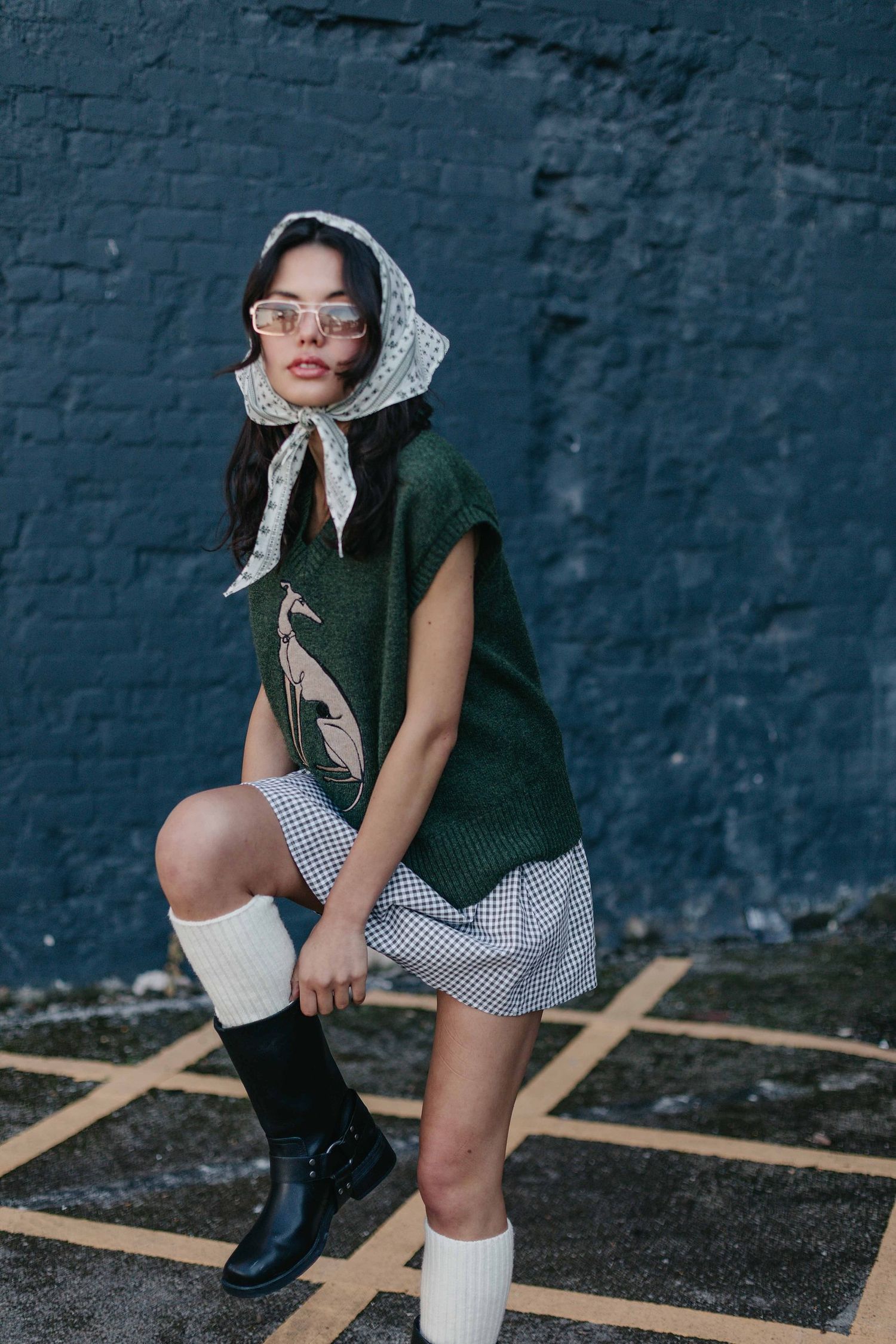 Woman in a patterned headscarf, green knit vest, checkered skirt and black boots poses against a dark blue brick wall.