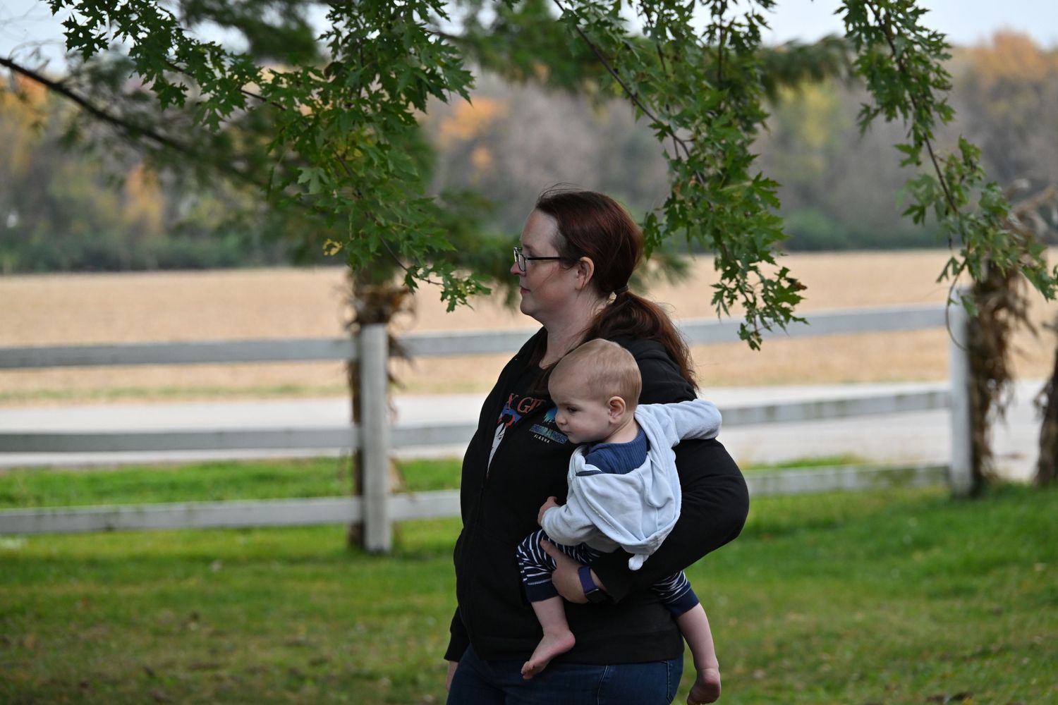 Woman with glasses and a dark jacket holds a baby in a white outfit while standing on a green lawn near a wooden fence and trees.