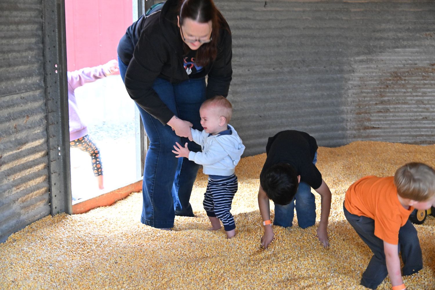A woman steadies a toddler standing in a barn filled with golden corn kernels while two other children play in the grain nearby.