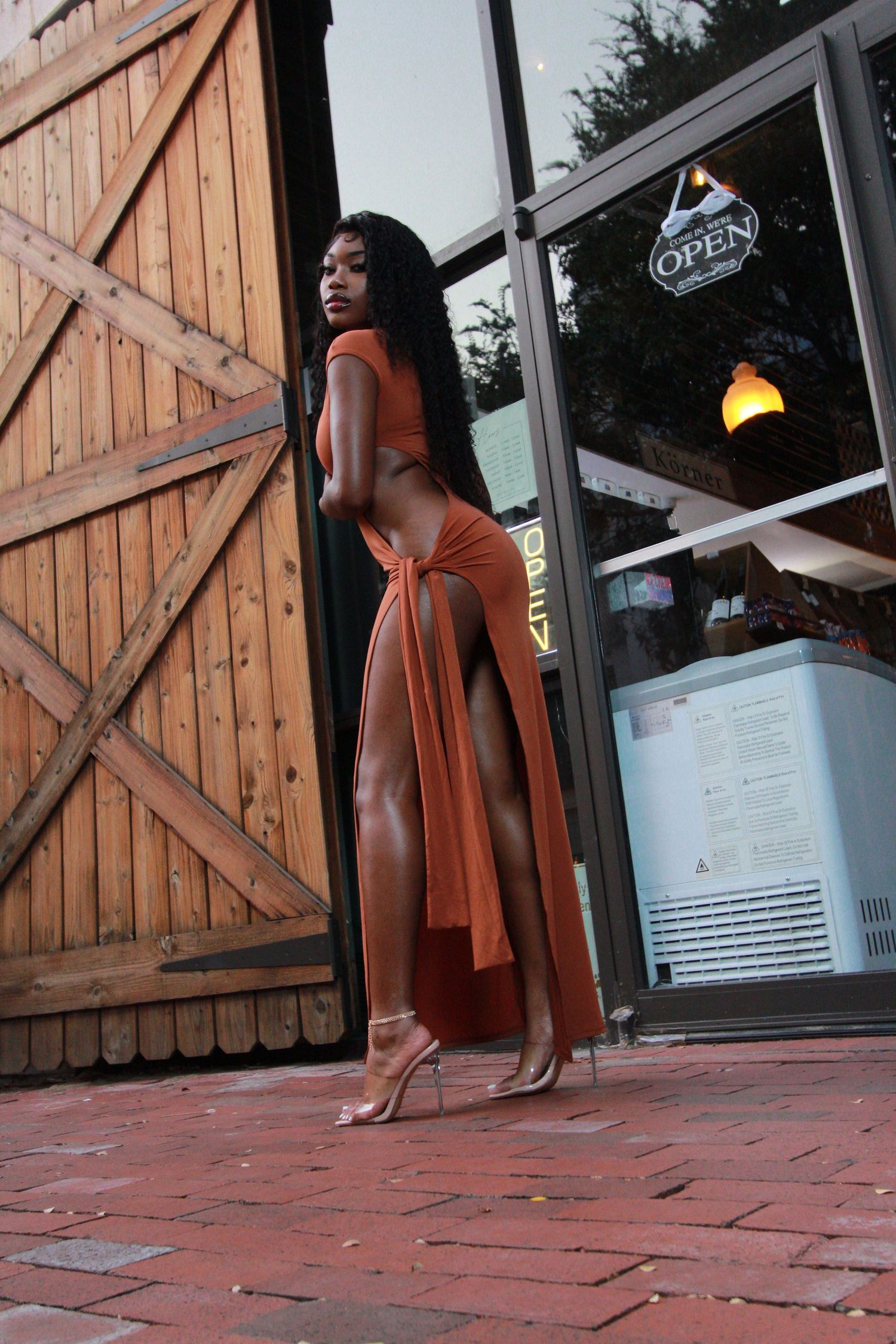A person in a flowing rust-colored dress poses elegantly against a wooden door outside a storefront with glowing lights.