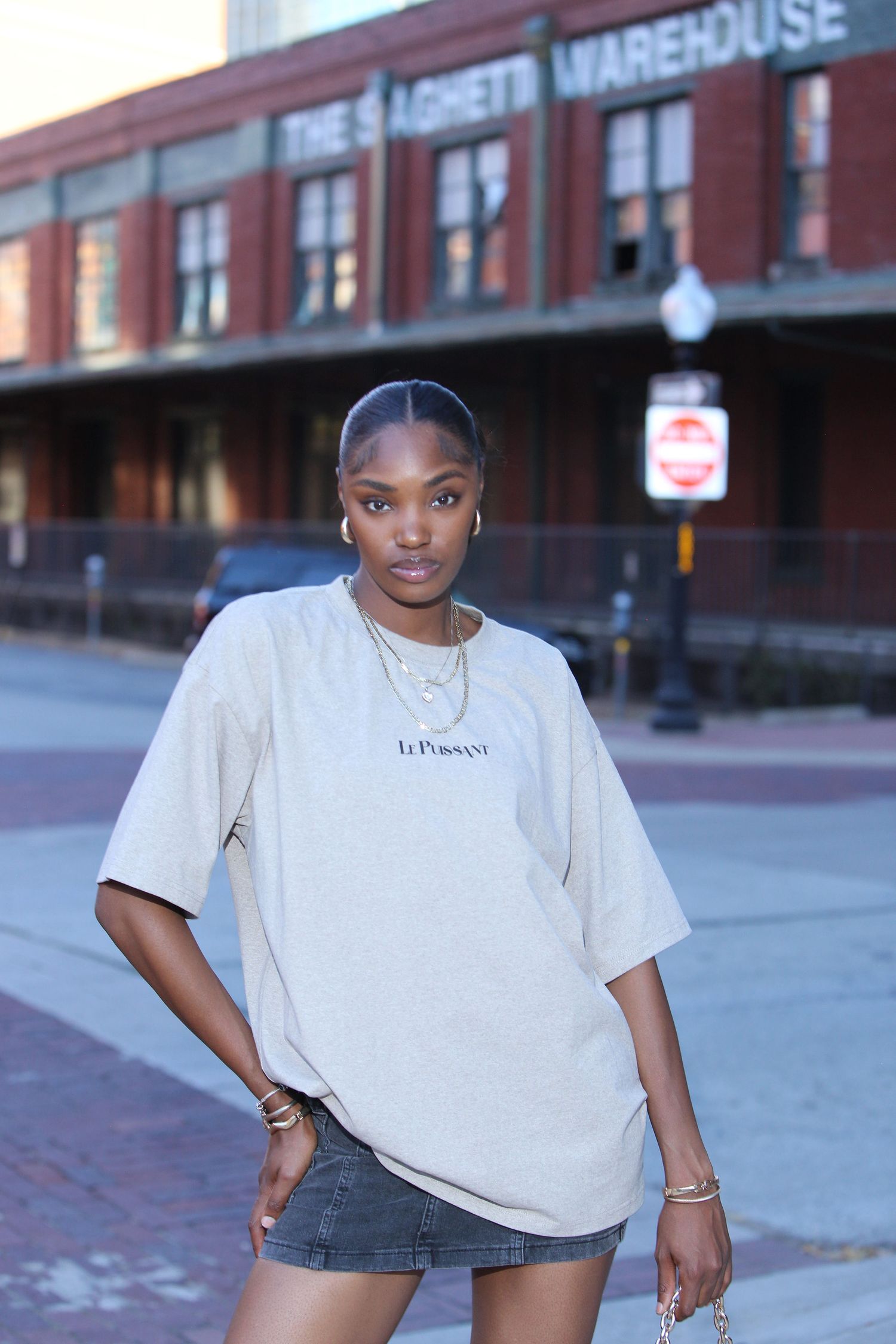 A person in an oversized beige t-shirt and denim shorts poses in front of The Barrel Warehouse building.