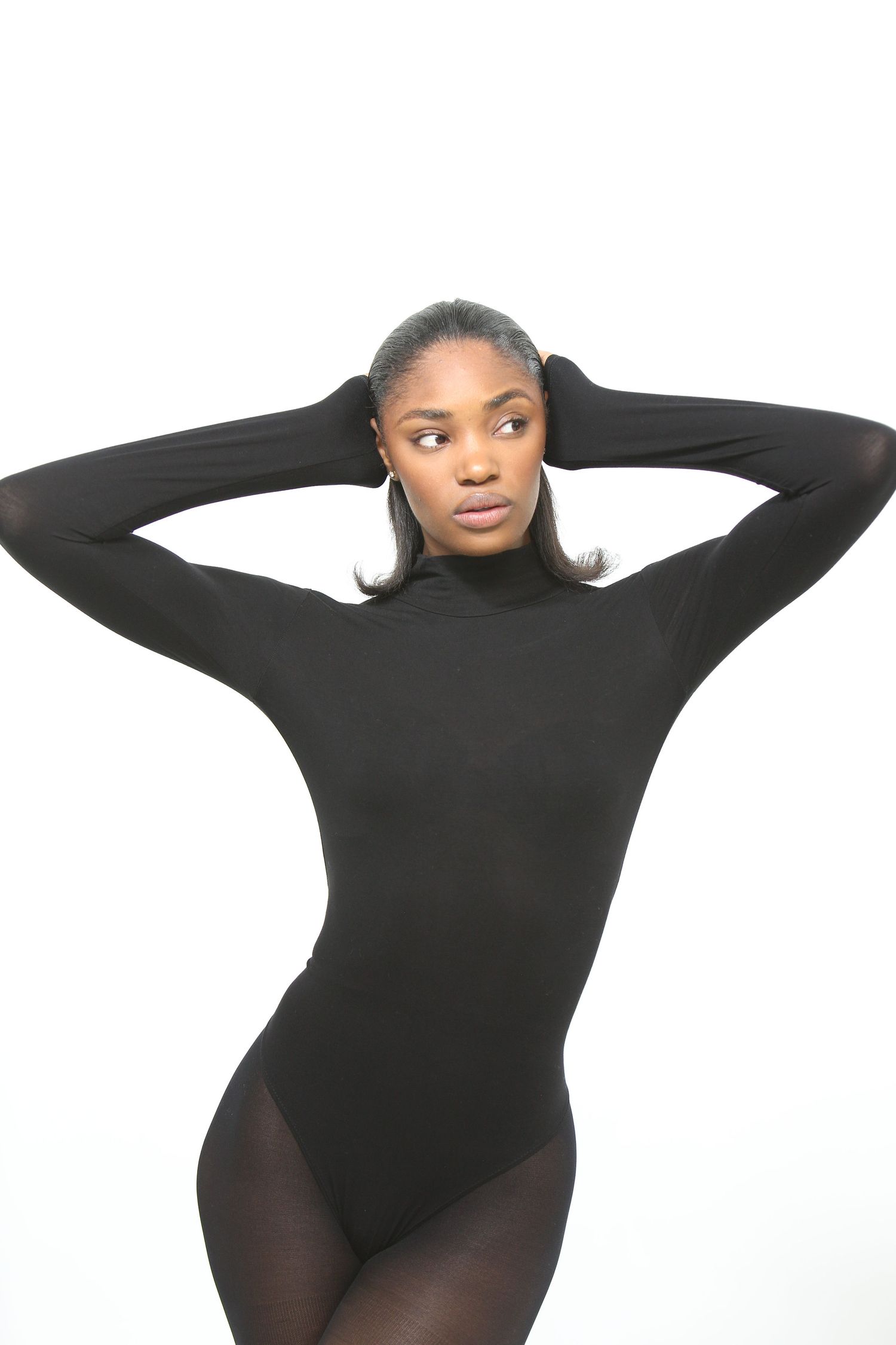 Black woman in a black long-sleeve turtleneck bodysuit and tights, posing with hands behind head on white background.