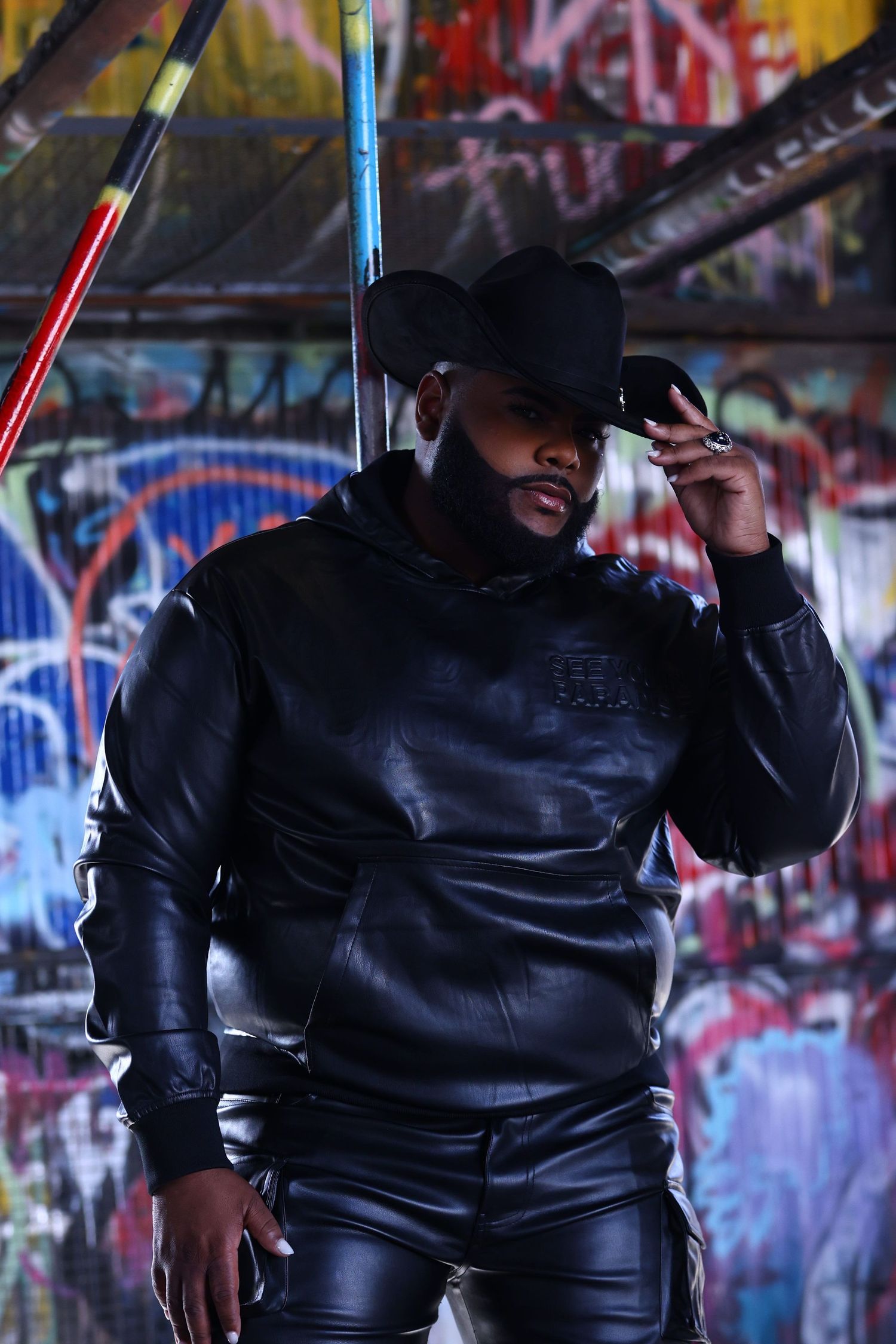 Man in all-black leather outfit and cowboy hat posing against colorful graffiti wall.