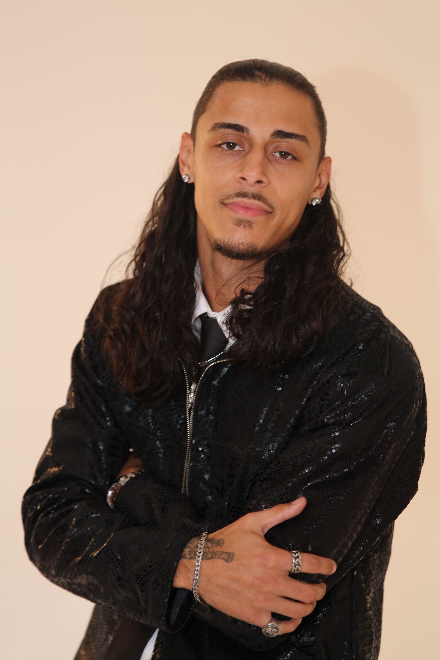 Young man with long dark hair wearing a black sequined jacket, arms crossed, posing against a beige background.