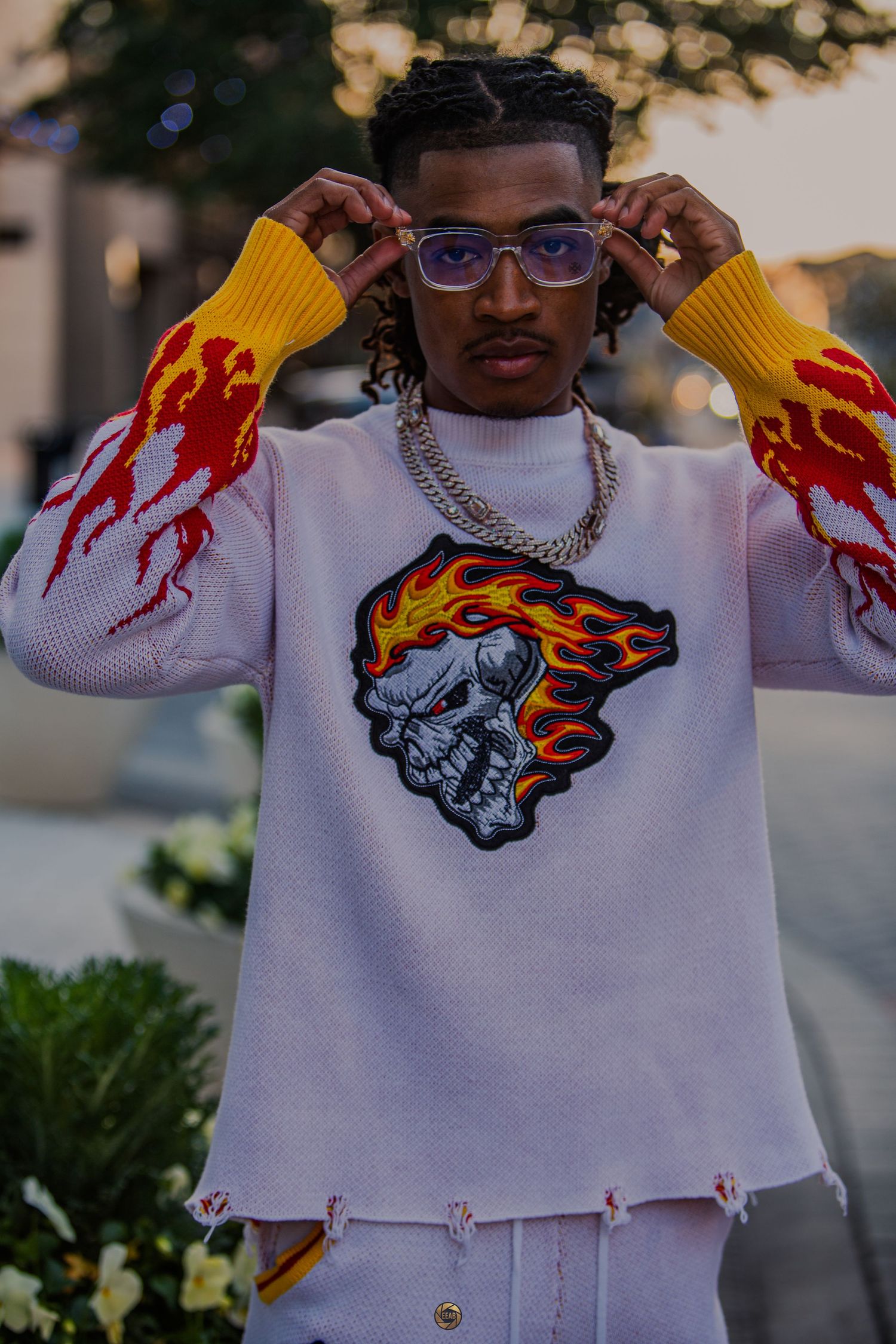 Young man wearing a flame-print sweater with skull graphic, silver chain necklace, and purple glasses posing outdoors.