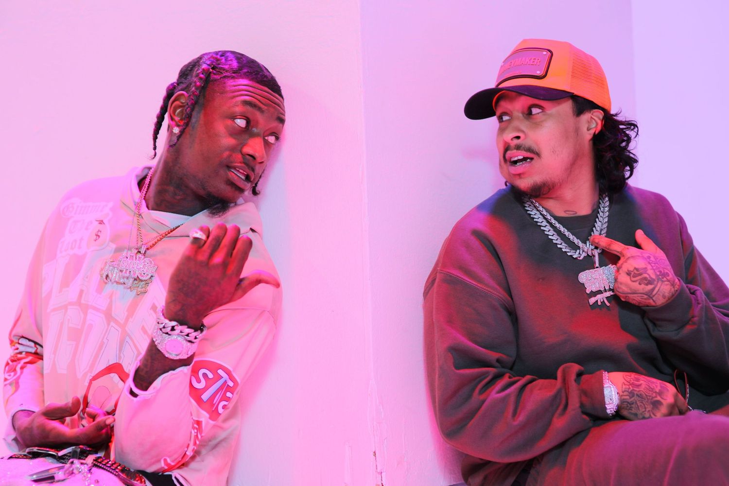 Two performers sit together against a pink-lit background wearing casual streetwear and making hand gestures Turnmeuptd