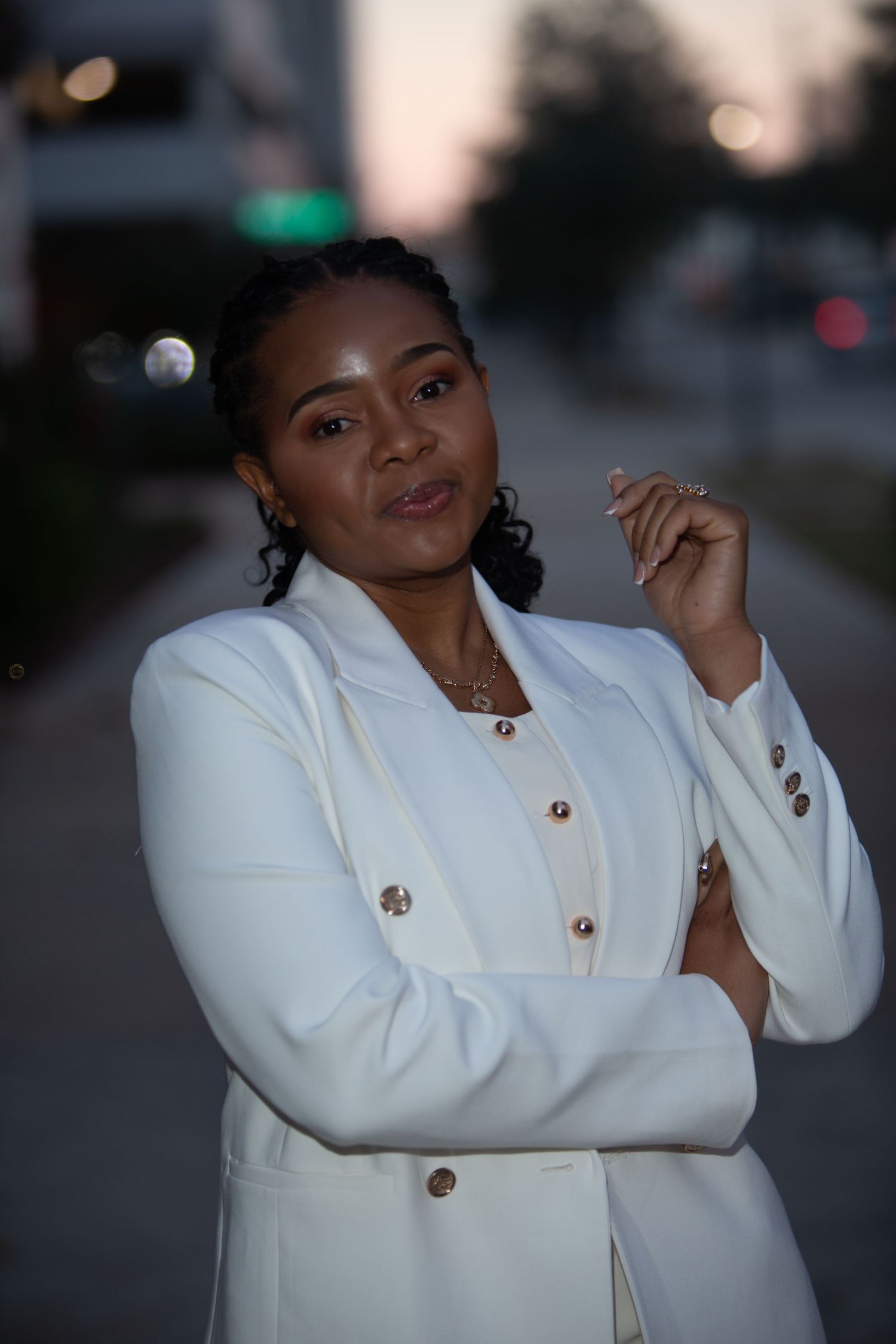A confident Black woman in a white blazer poses outdoors at dusk with arms crossed, wearing gold jewelry.