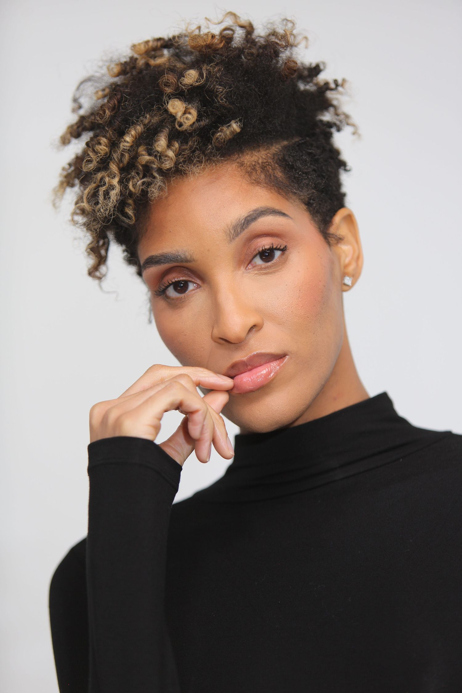 Young woman with curly updo hairstyle wearing black turtleneck, posing thoughtfully with hand near chin.