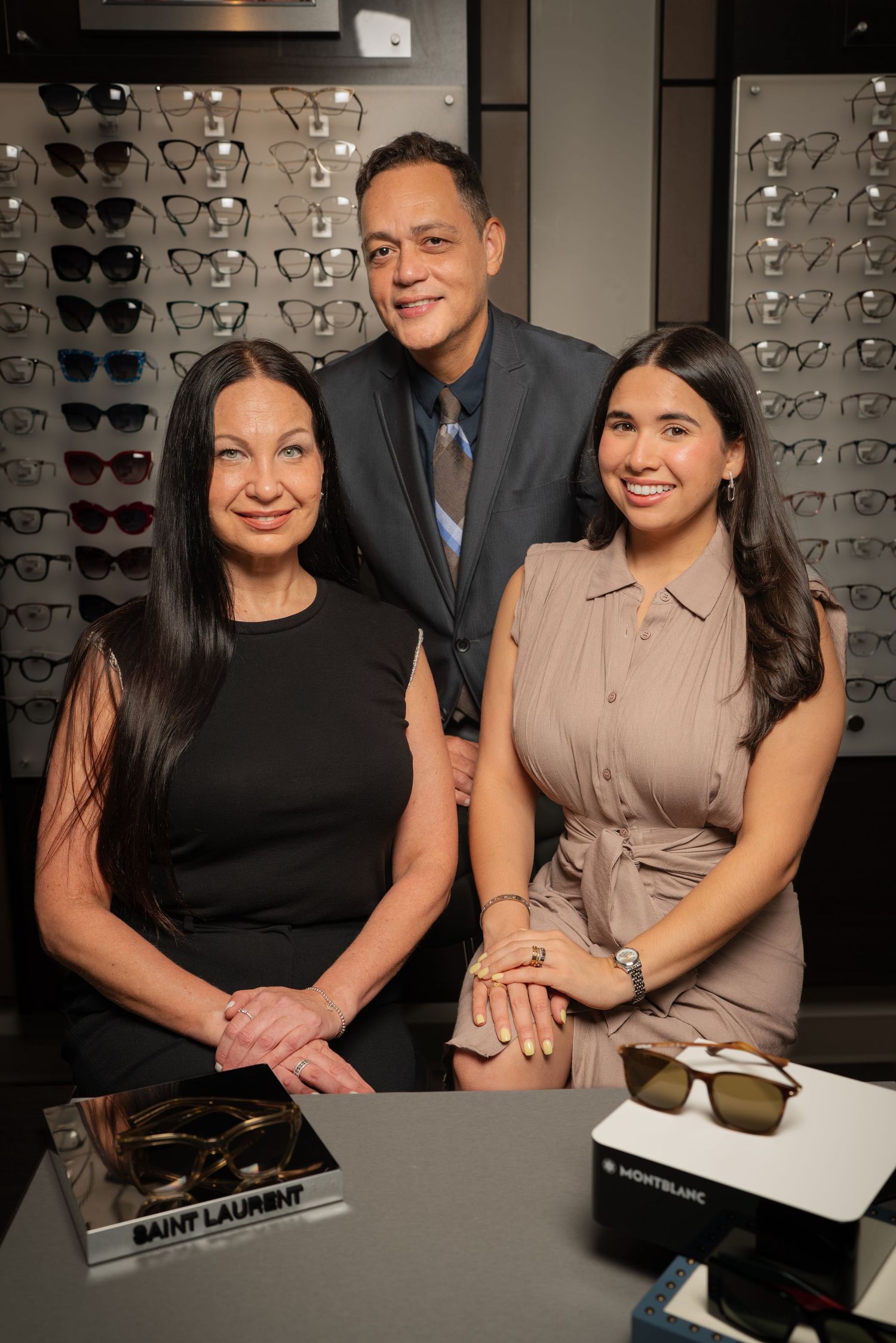 Three smiling optometry staff pose in an eyewear store with Saint Laurent and Montblanc glasses on display.