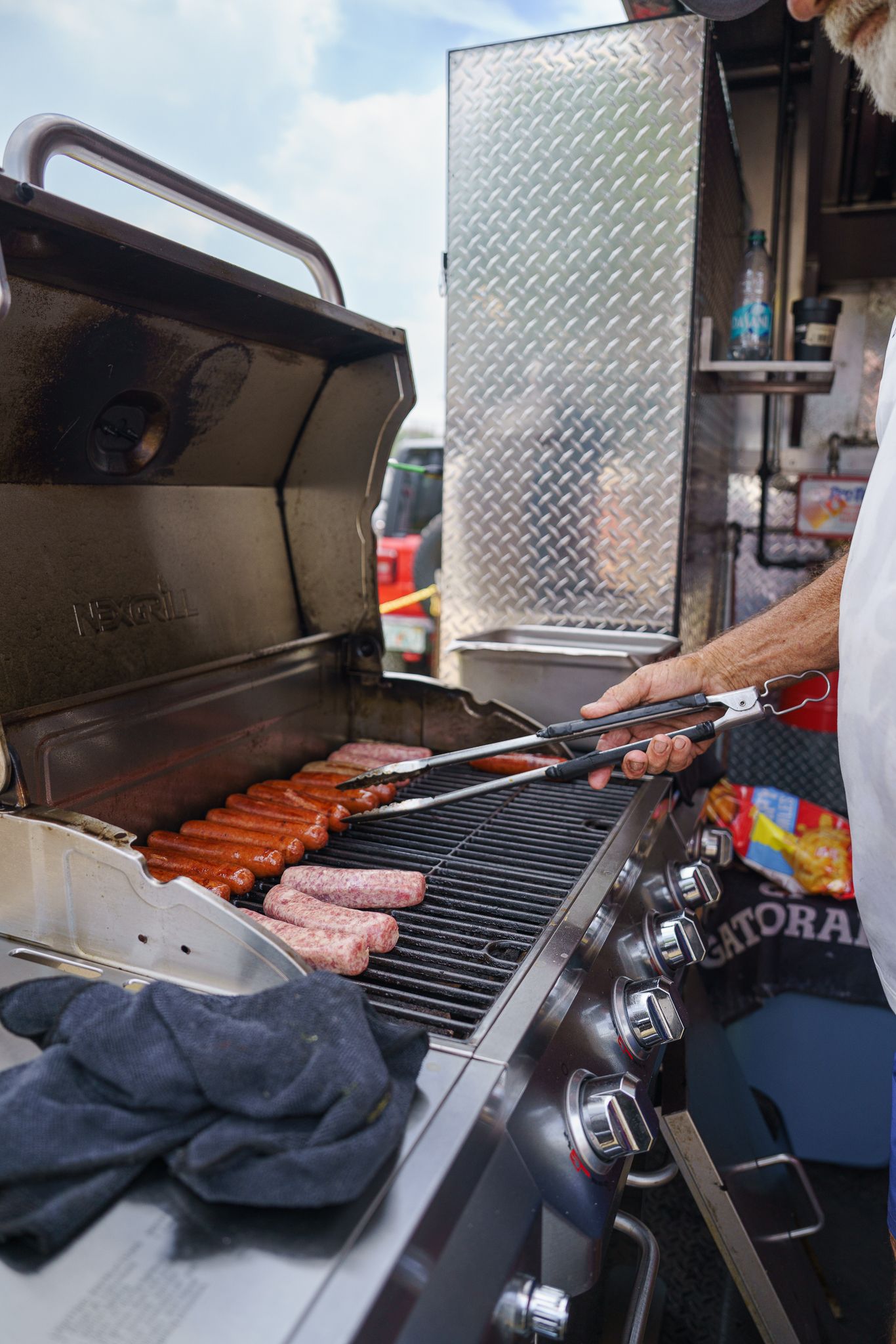 Person grilling hot dogs and sausages on a gas grill at an outdoor food vendor stand.