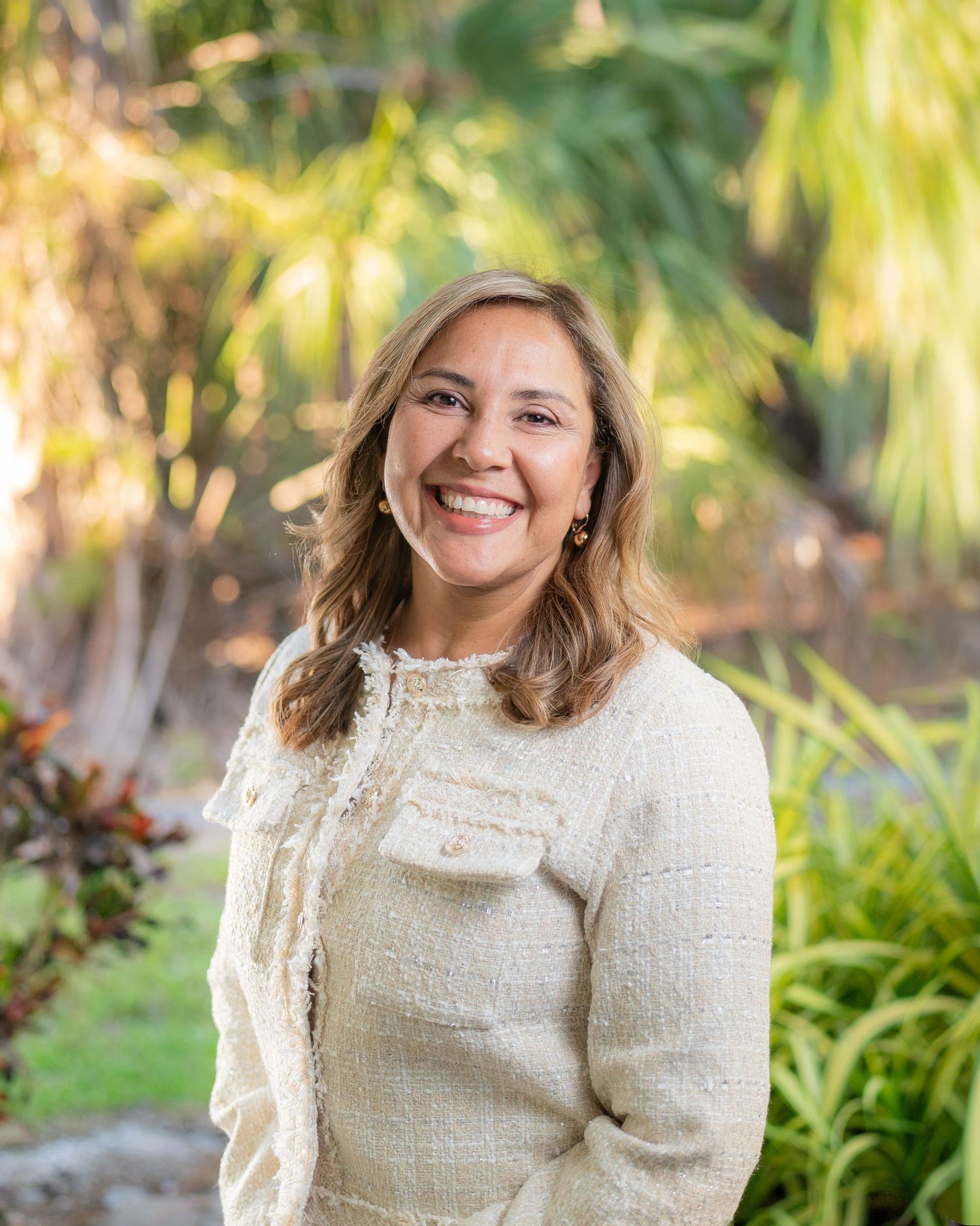 Smiling woman with wavy hair wearing cream tweed jacket poses outdoors among lush tropical greenery.