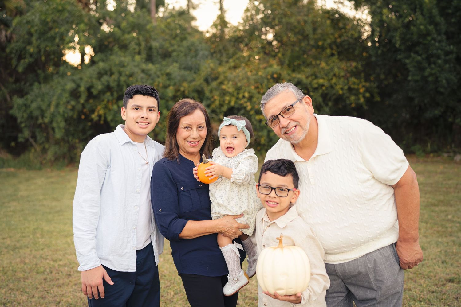 Multigenerational family of five posing outdoors with a baby and white pumpkin at golden hour.