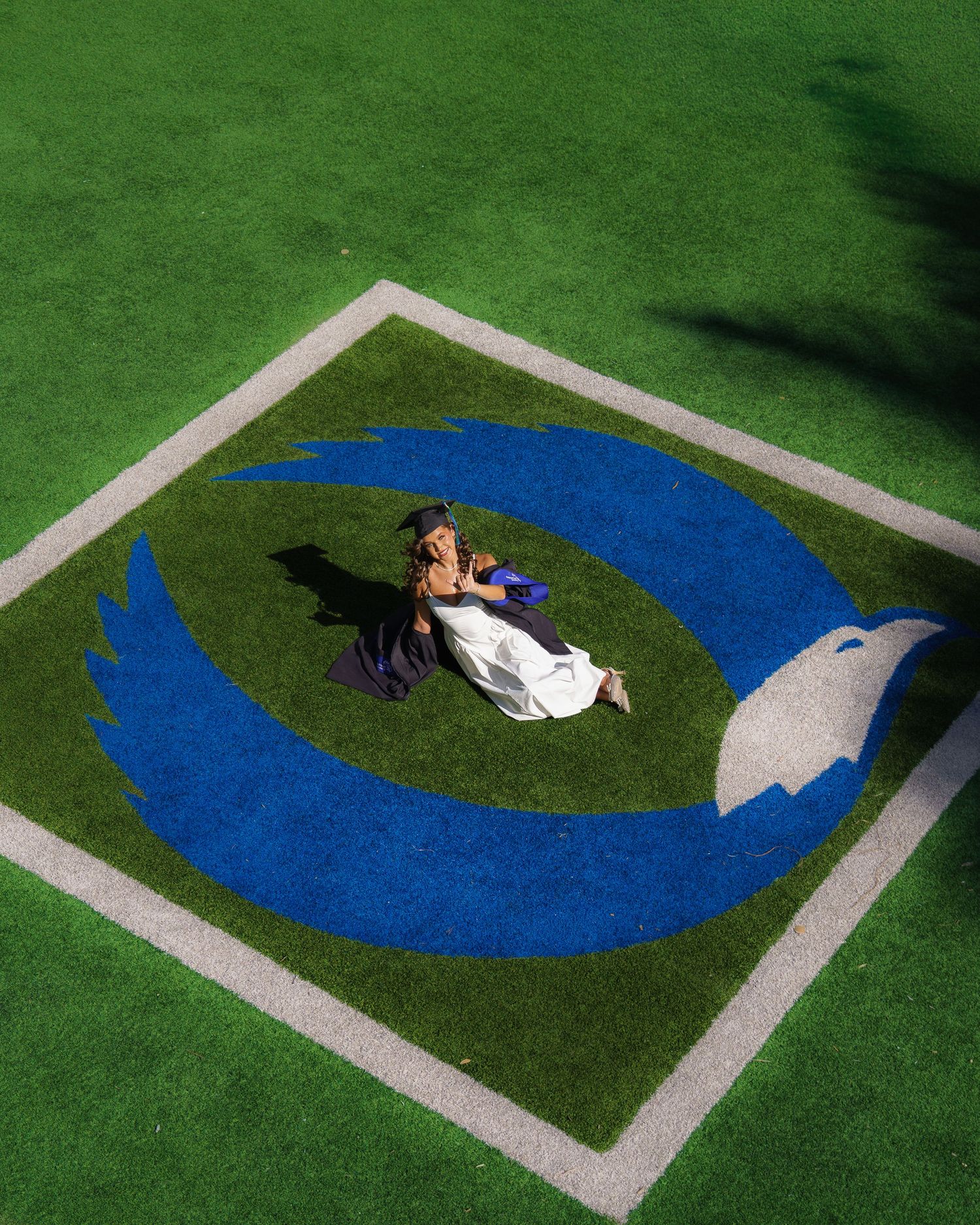 Aerial view of two graduates sitting on green turf featuring a large blue bird logo painted on the field.