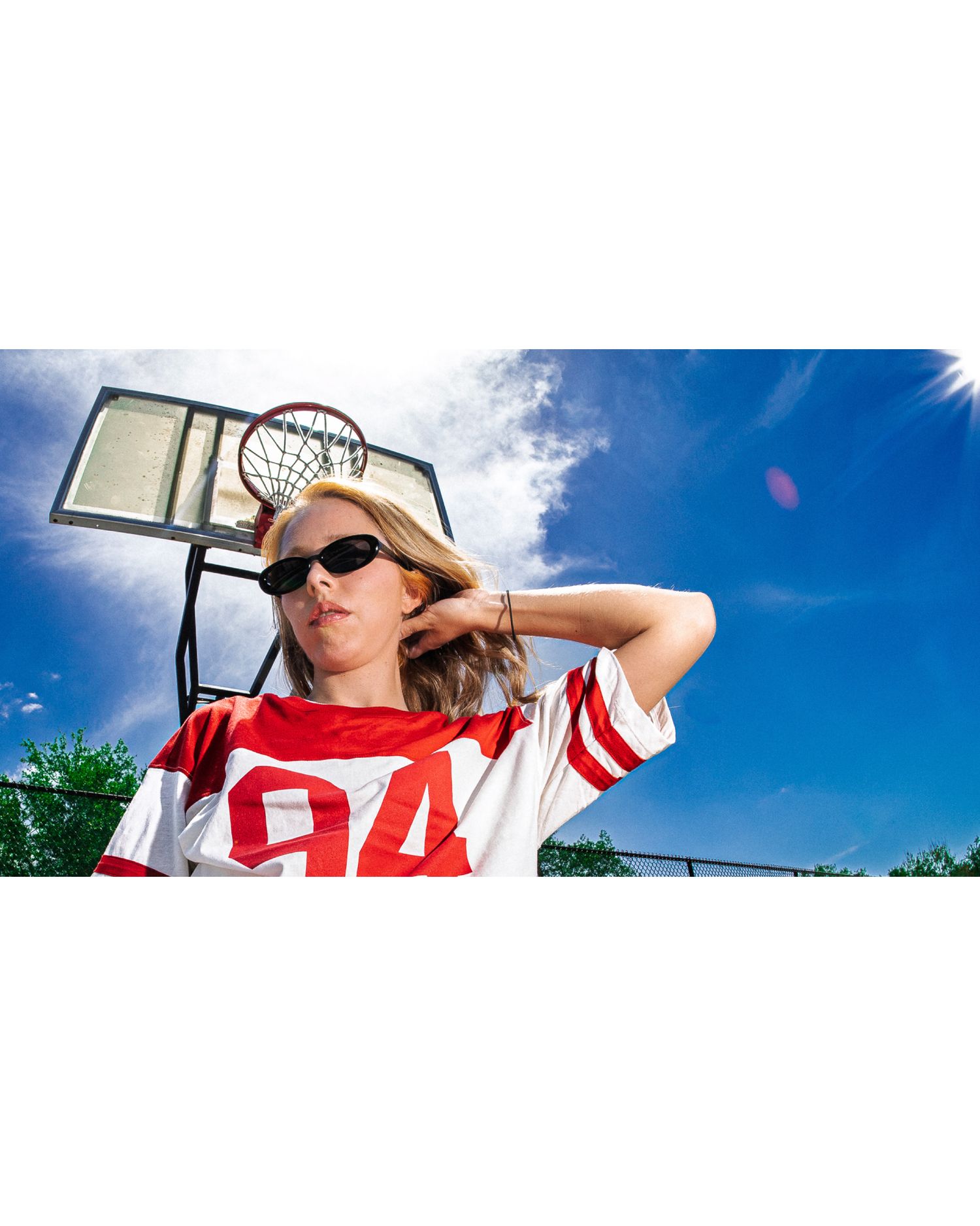 Young woman in red and white jersey #94 posing at outdoor basketball court wearing sunglasses on sunny day.