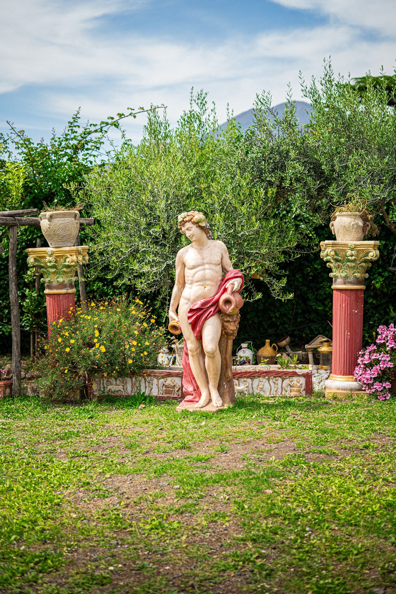 Classical Roman statue of a male figure draped in red cloth holding an amphora in a lush garden setting.