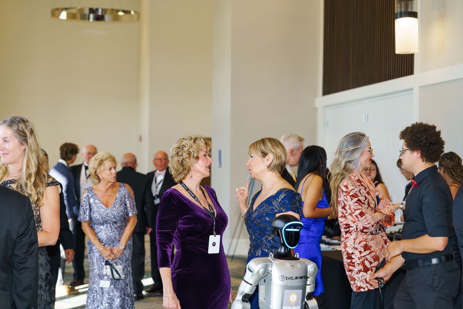 Elegantly dressed guests mingle at an upscale indoor event, with women in formal gowns socializing in a bright venue.
