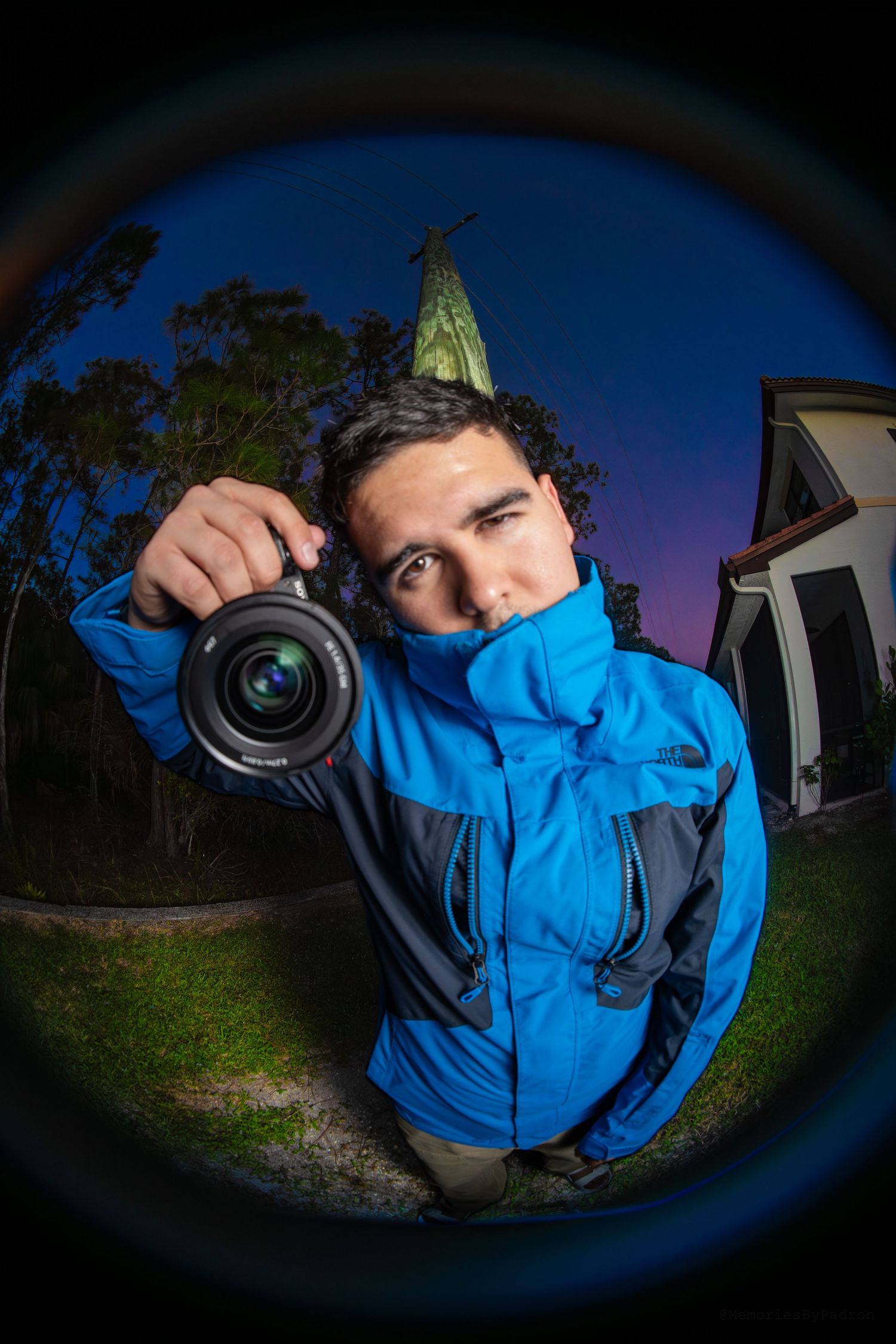 Photographer holding camera lens up in fisheye shot at night near illuminated statue outdoors.