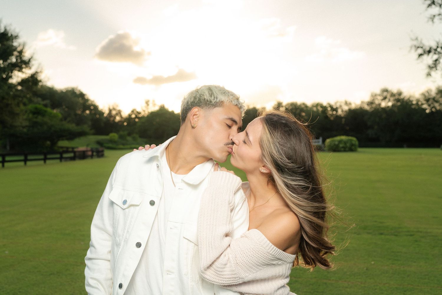 Romantic couple sharing a kiss on a green field at sunset with warm glowing sky.