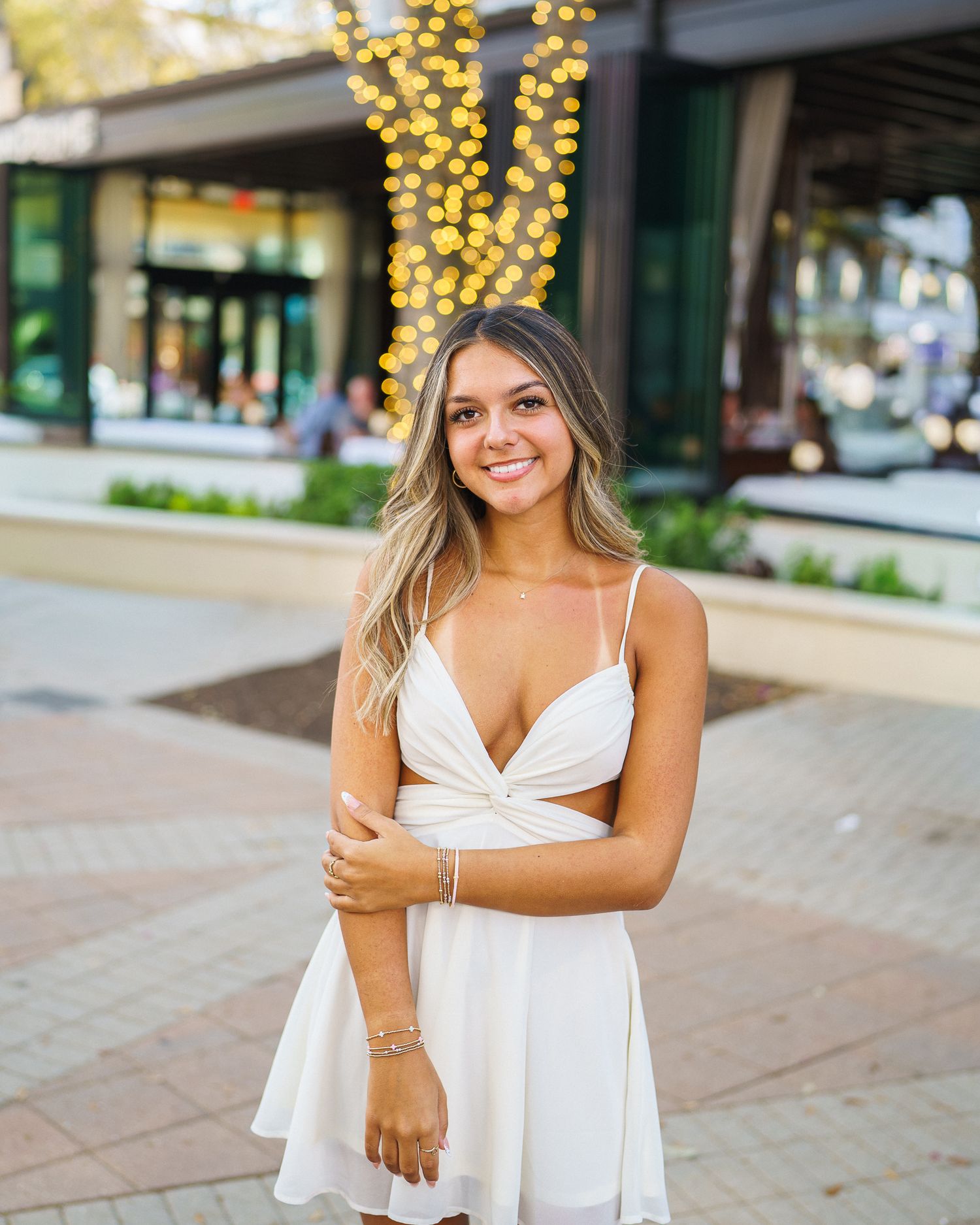Young woman in white mini dress smiling in front of bokeh string lights at outdoor venue.