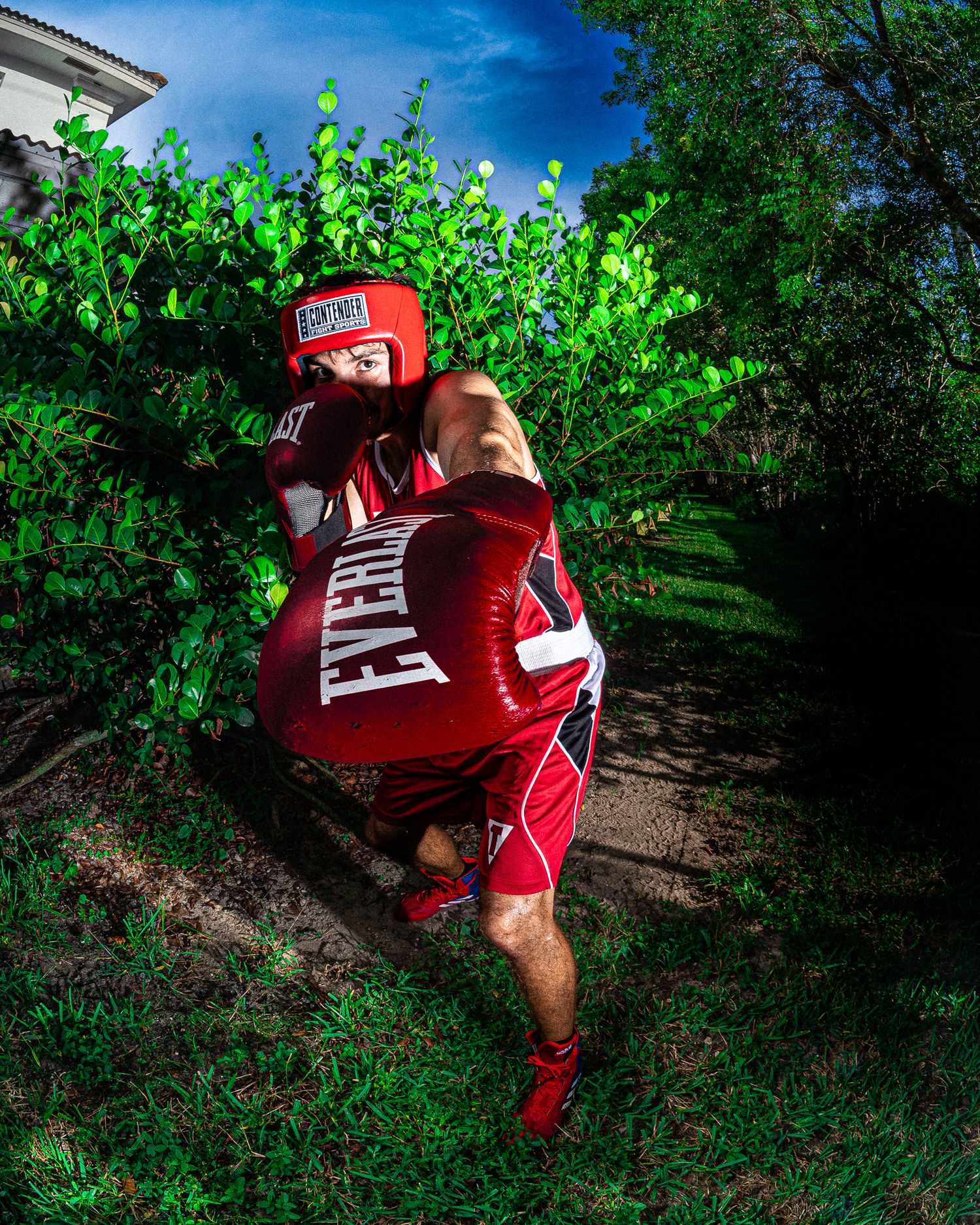 Boxer in red Everlast gear throwing a punch outdoors, wearing headgear and gloves near lush green bushes.