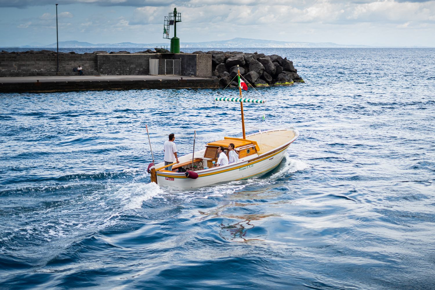 A small white and yellow wooden boat sailing near a stone pier with a green lighthouse in calm blue waters.
