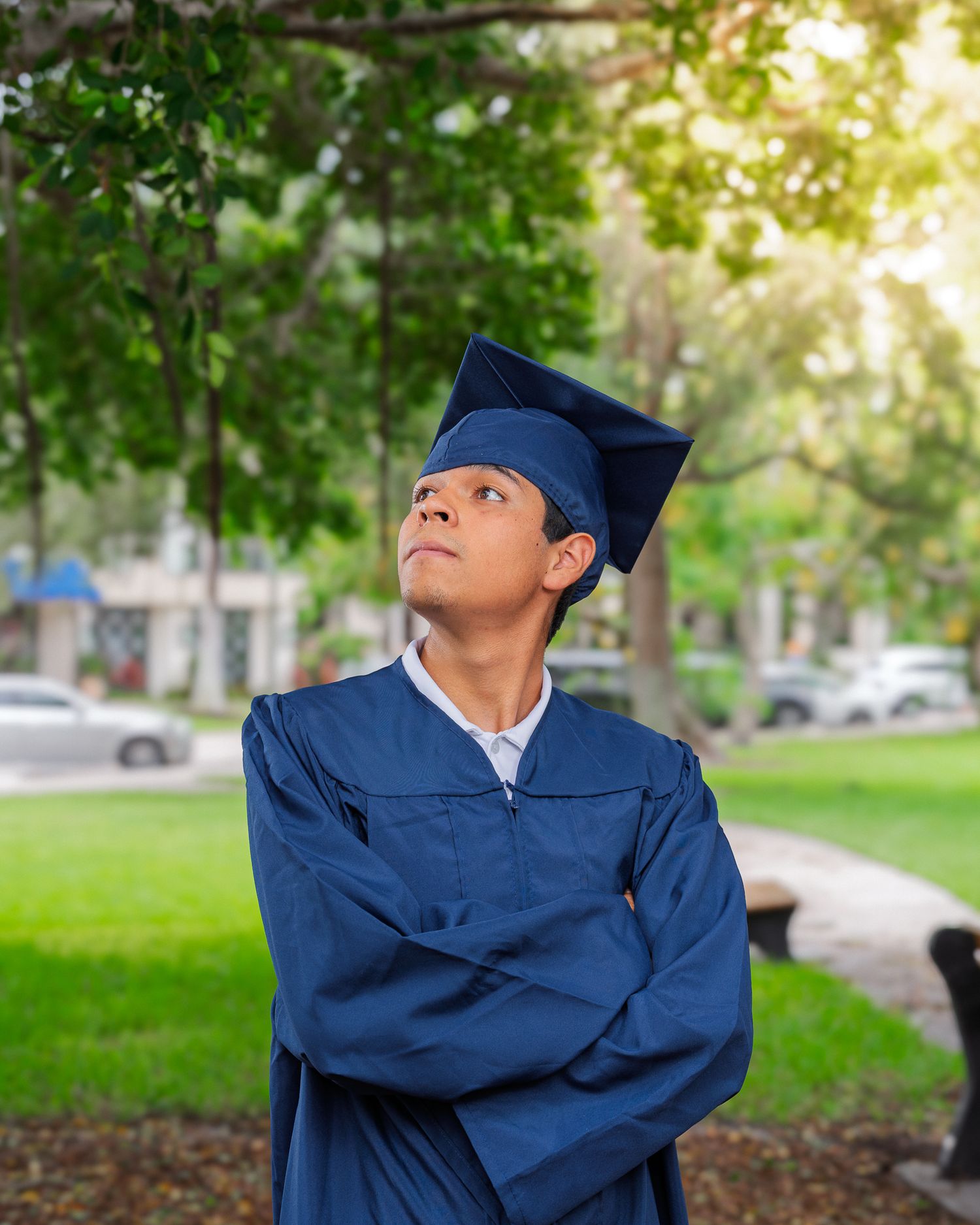 A young graduate in blue cap and gown stands outdoors with arms crossed, looking upward thoughtfully.