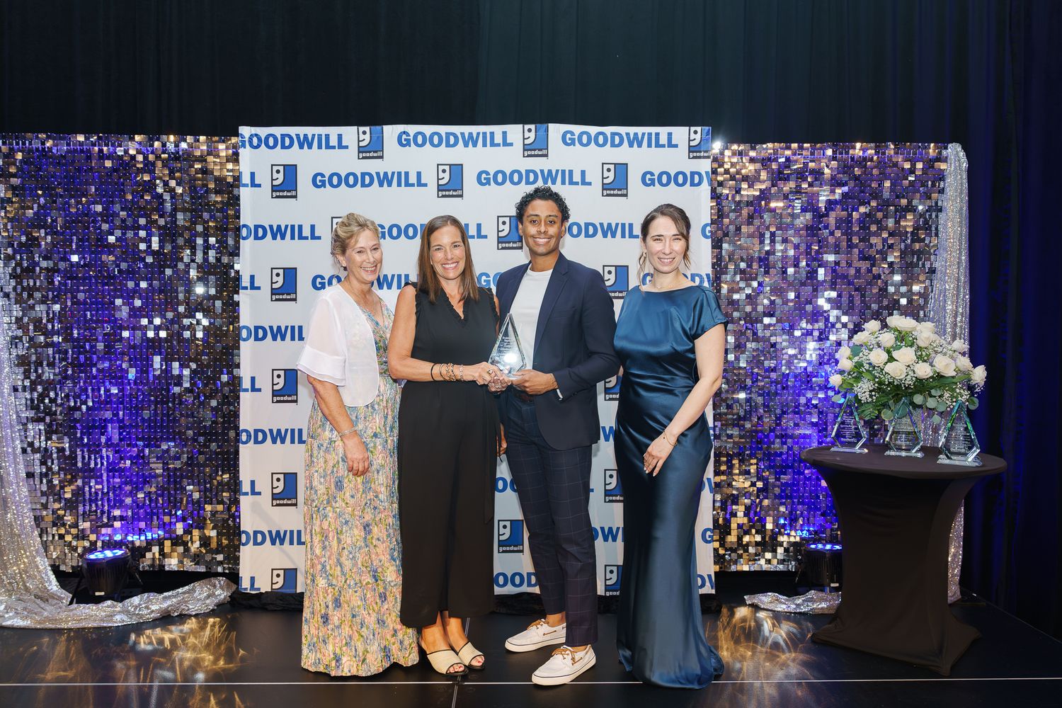 Four people pose together at a Goodwill awards ceremony in front of a sparkling blue sequin backdrop.
