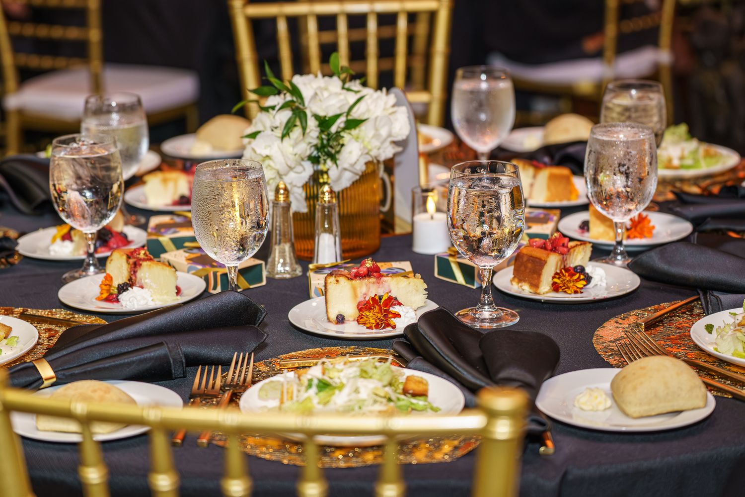 Elegant banquet table set with gold chairs, black linens, white floral centerpiece, and plated appetizers with wine glasses.