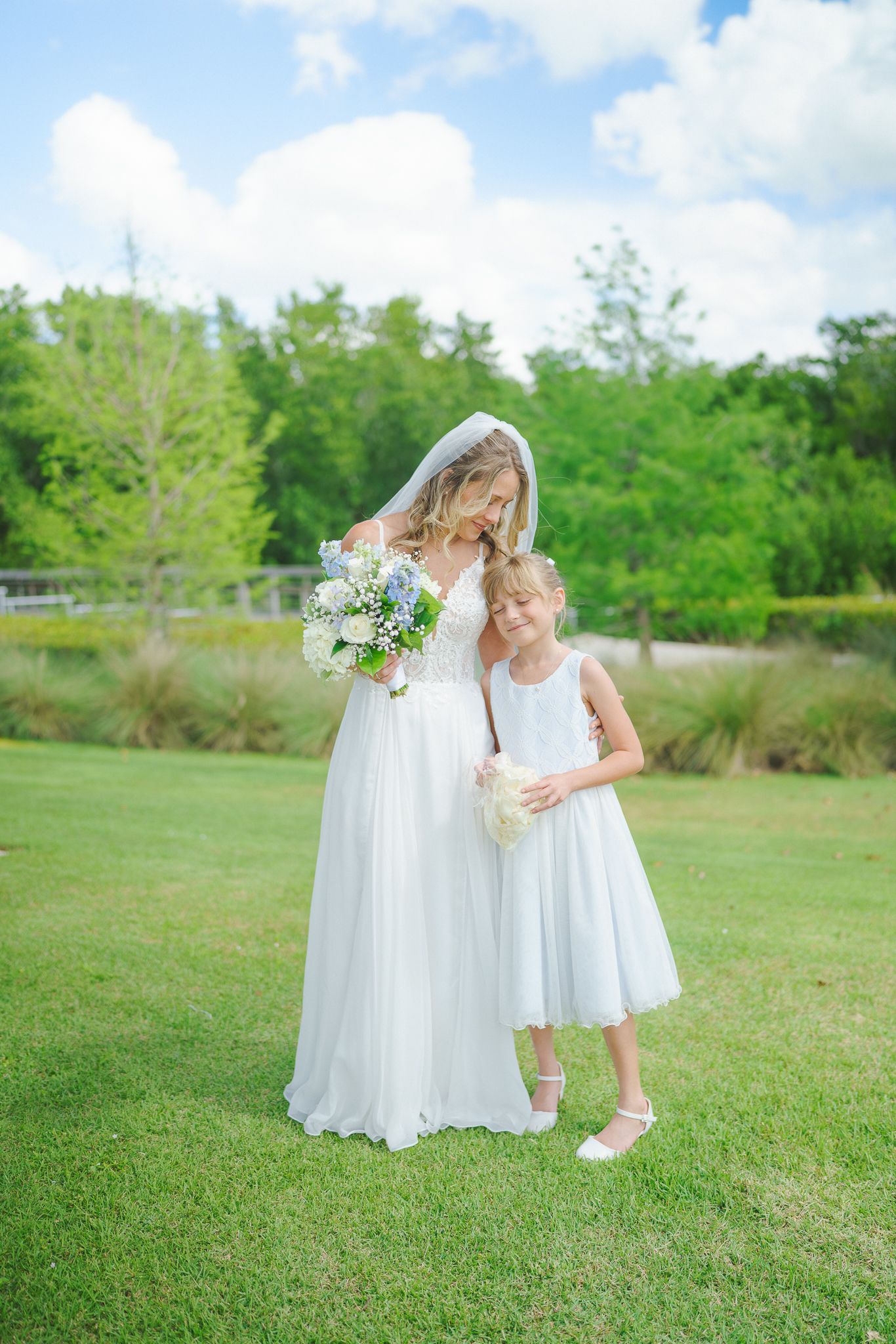 Bride in white gown and veil poses with young flower girl on green lawn outdoors.