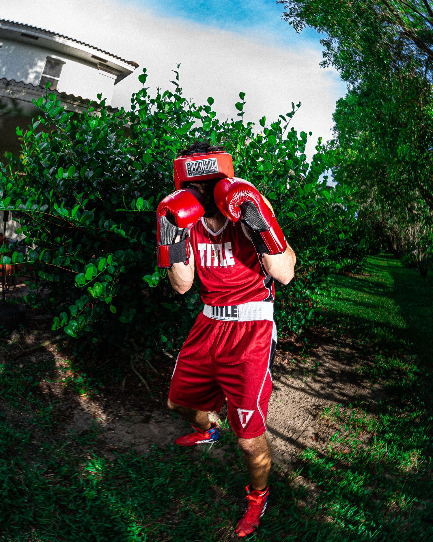 A boxer in red Title boxing gear and gloves poses in a defensive stance outdoors among lush green trees.