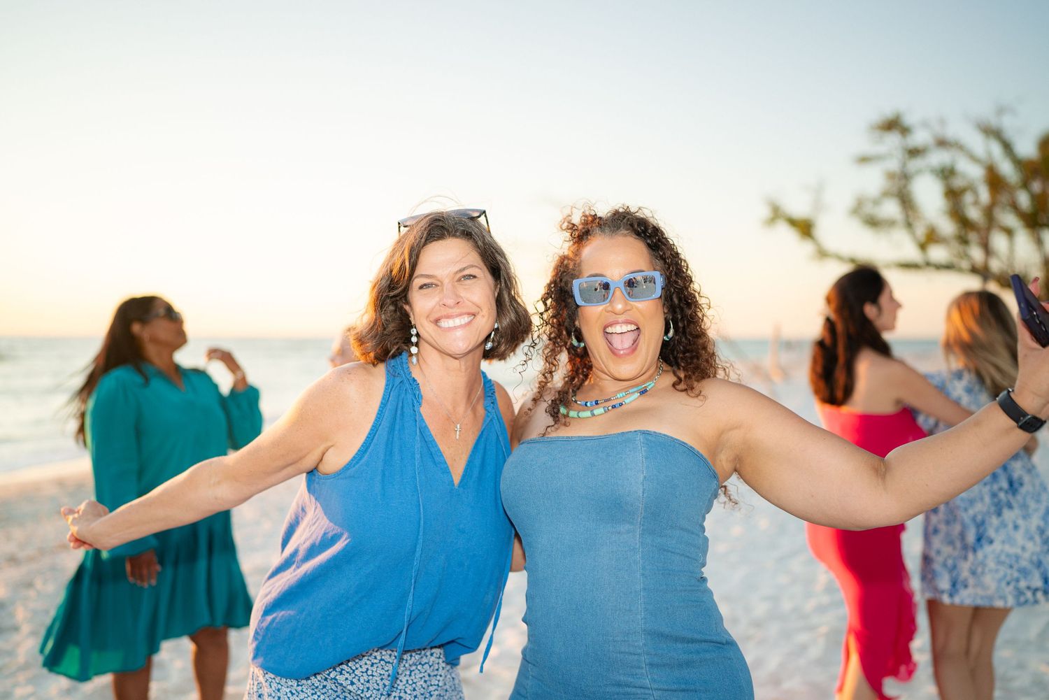 Two smiling women in blue outfits posing together at a beach party during sunset.